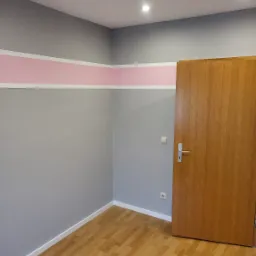 Pokój po remoncie: szare ściany z poziomym pasem w kolorze pudrowego różu, drewniane panele podłogowe, brązowe drzwi z srebrną klamką, białe listwy przypodłogowe.