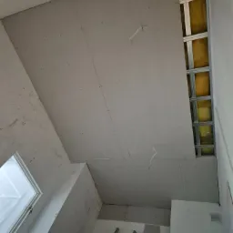 Montaż sufitu podwieszanego z płyt karton-gips, widoczne profile metalowe, izolacja z wełny mineralnej oraz częściowo zamontowane płyty. W tle drabina i okno.