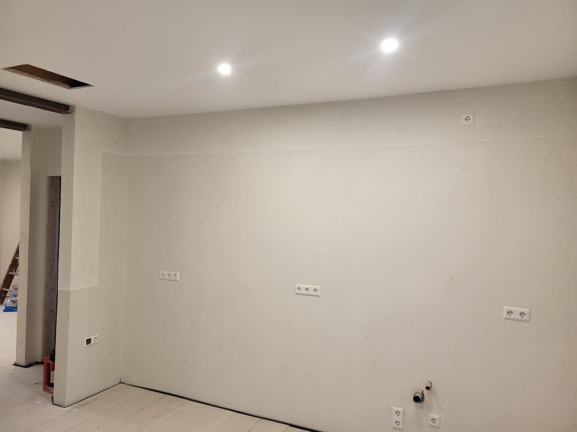 Świeżo pomalowane wnętrze z widocznymi gniazdkami elektrycznymi i instalacją hydrauliczną, sufit z otworem po demontażu oświetlenia, podłoga zabezpieczona przed zabrudzeniem.