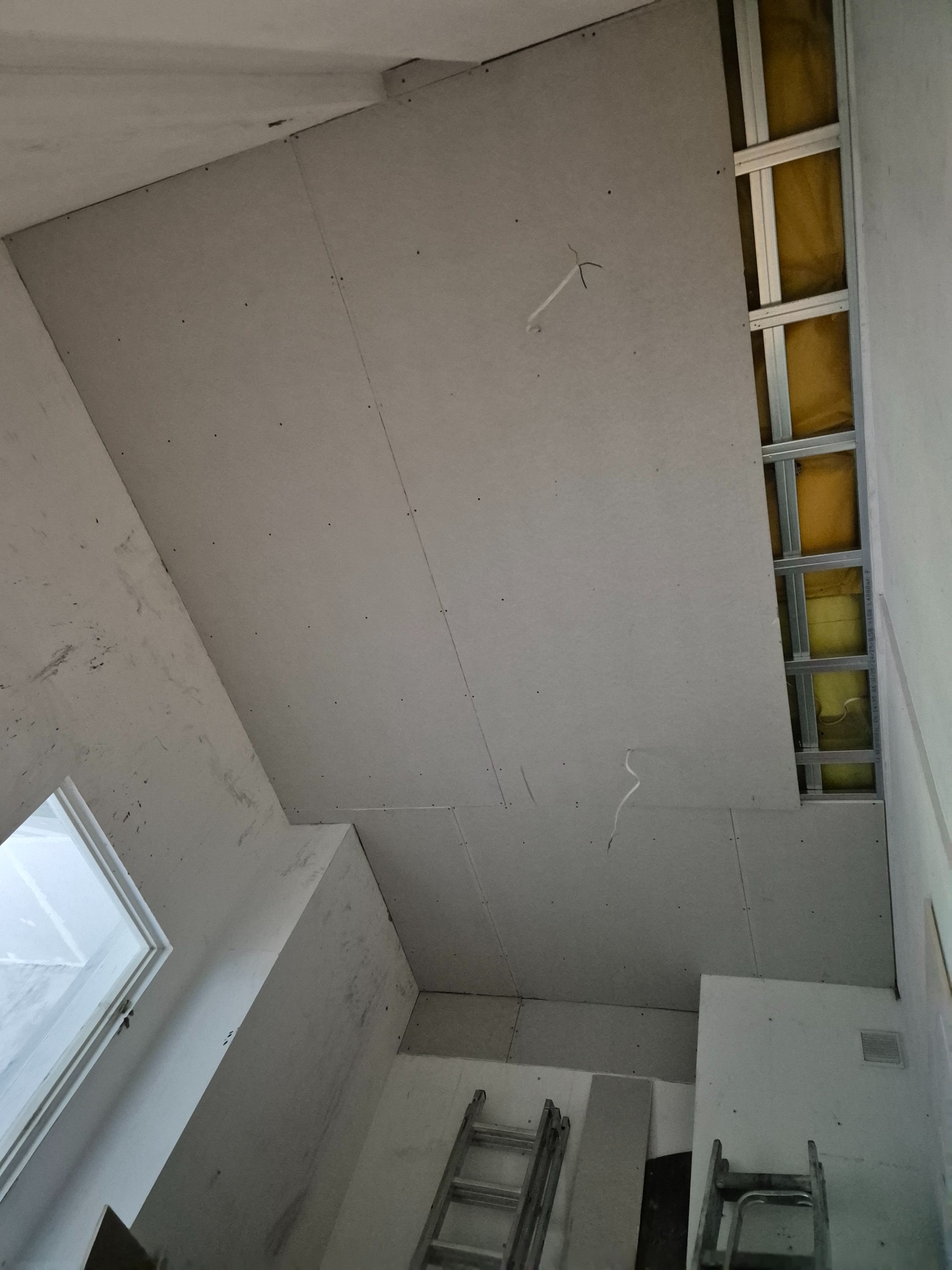 Montaż sufitu podwieszanego z płyt karton-gips, widoczne profile metalowe, izolacja z wełny mineralnej oraz częściowo zamontowane płyty. W tle drabina i okno.