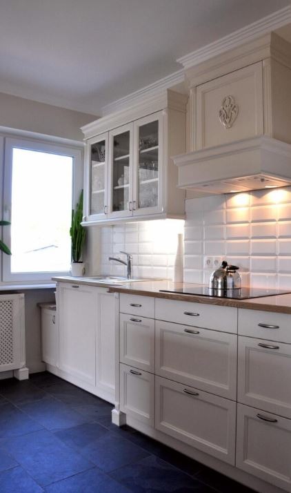 Jasna kuchnia z białymi szafkami z frezowanymi frontami, szklanymi witrynami górnymi i drewnianym blatem, uzupełniona o białe płytki ścienne i czarną podłogę.