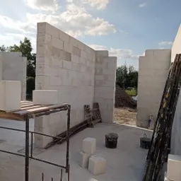 Wnętrze wznoszonego domu z białych bloczków, widoczne fragmenty ścian, rusztowanie, drabina oparta o ścianę i wiadro. W tle zieleń drzew i niebo z chmurami.