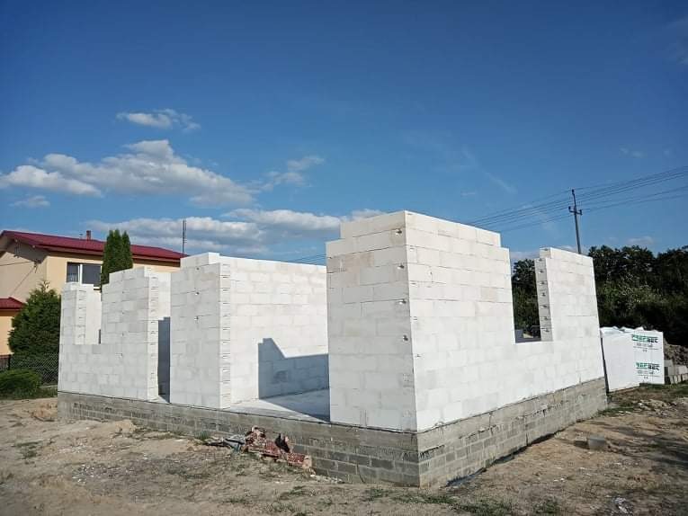 Wznoszenie ścian parterowego domu z białych bloczków na fundamencie z cegły, widok na budynek w stanie surowym otwartym, błękitne niebo z obłokami w tle.
