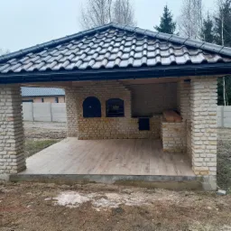 Zadaszona altana z cegły z czarną dachówką ceramiczną, drewnianą podłogą i murowanymi filarami, na zewnątrz w pochmurny dzień.