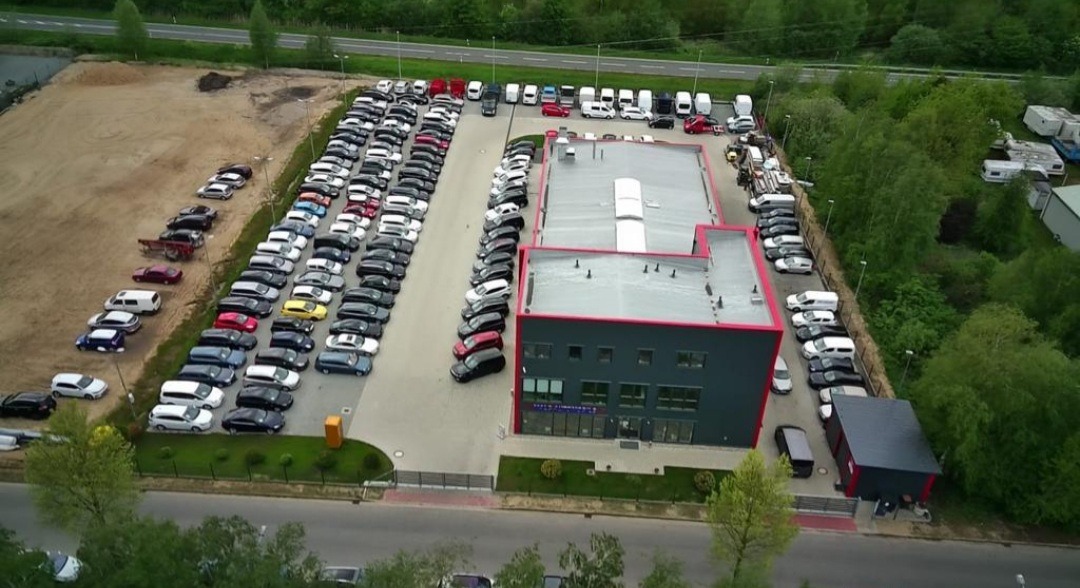 Widok z drona na parking pełen samochodów osobowych i dostawczych, z budynkiem biurowym w tle, otoczonym zielenią.