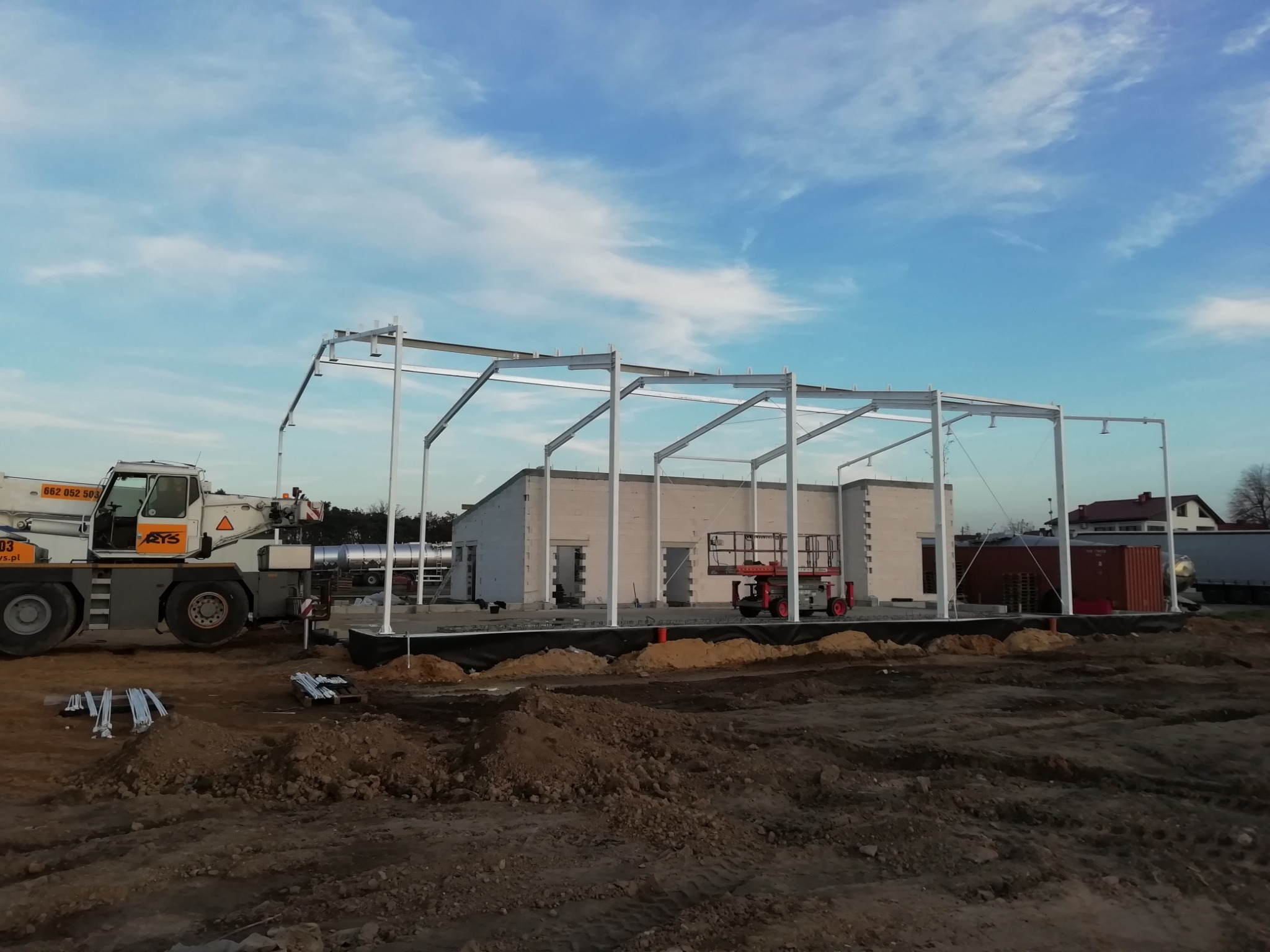Montaż stalowej konstrukcji hali magazynowej z użyciem dźwigu teleskopowego; widoczne fundamenty, w tle budynek w trakcie budowy i błękitne niebo.