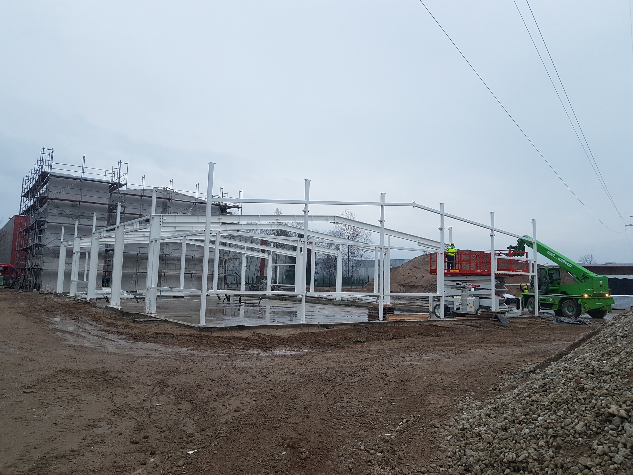 Montaż stalowej konstrukcji hali magazynowej z wykorzystaniem podnośnika koszowego i żurawia teleskopowego; w tle budynek w rusztowaniach.