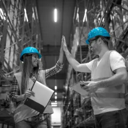 Dwoje pracowników magazynu w niebieskich kaskach i kamizelkach odblaskowych przybija piątkę na tle wysokich regałów wypełnionych towarami, zdjęcie w czerni i bieli z akcentem na niebieskie kaski.