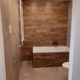 Wykończona łazienka z drewnopodobnymi płytkami na ścianie za wanną, szarymi płytkami na pozostałych ścianach i podłodze, białą wanną i grzejnikiem drabinkowym, oraz widoczną częścią toalety po...