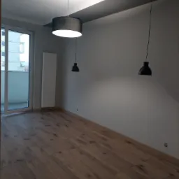 Pusty pokój po remoncie z drewnianą podłogą, szarą ścianą i trzema lampami wiszącymi: jedną okrągłą nad głową i dwiema mniejszymi, czarnymi po bokach. Na ścianie widoczny biały grzejnik.