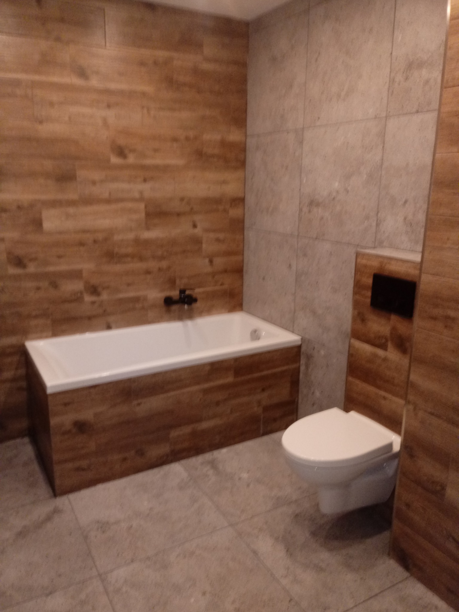 Wykończona łazienka z wanną obudowaną panelami imitującymi drewno, wiszącą toaletą i płytkami w odcieniach beżu na ścianach.