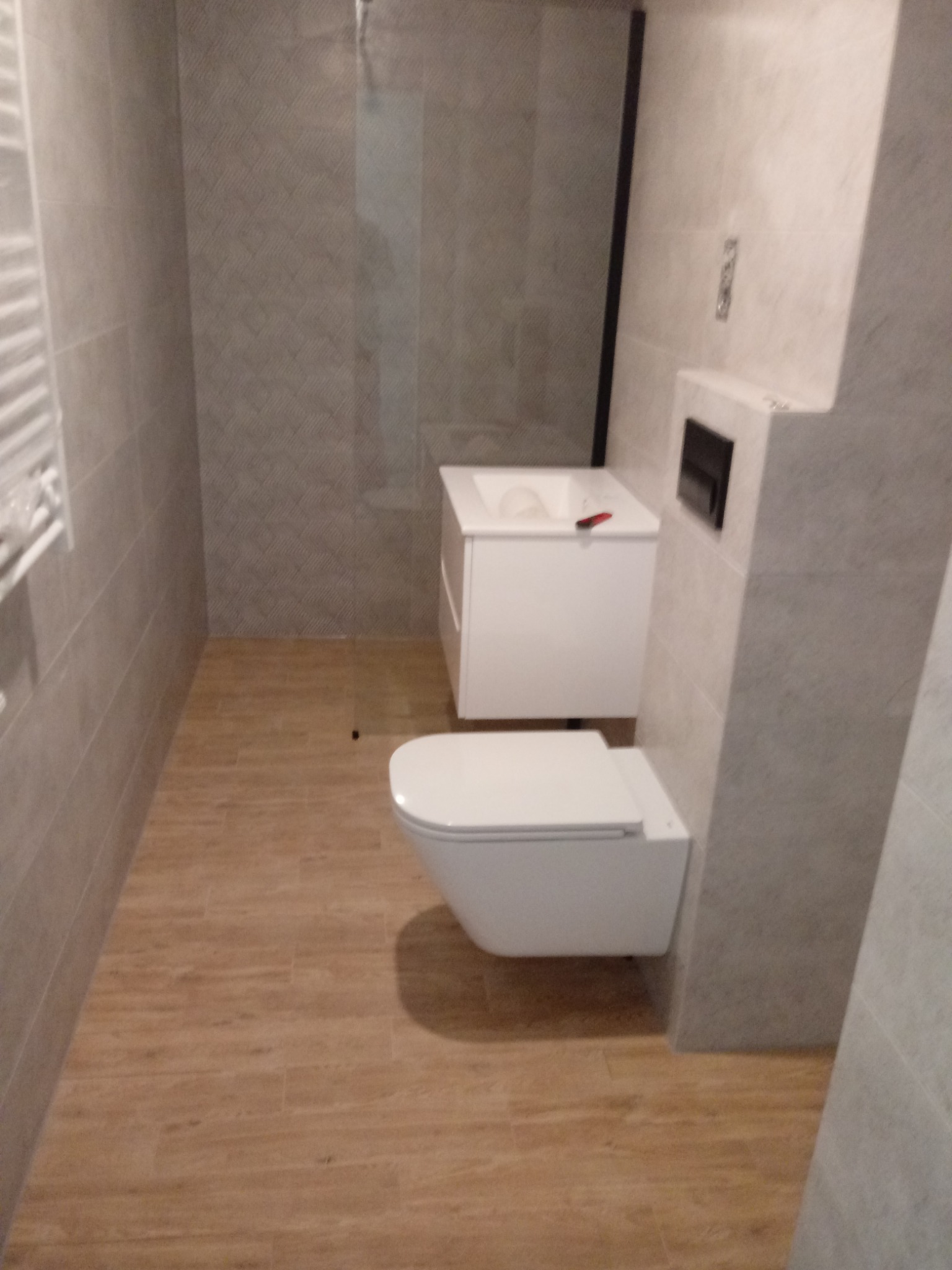 Nowoczesna łazienka z beżowo-szarą glazurą, drewnopodobną podłogą, wiszącą toaletą i białą szafką pod umywalką z czarną ramą lustra.