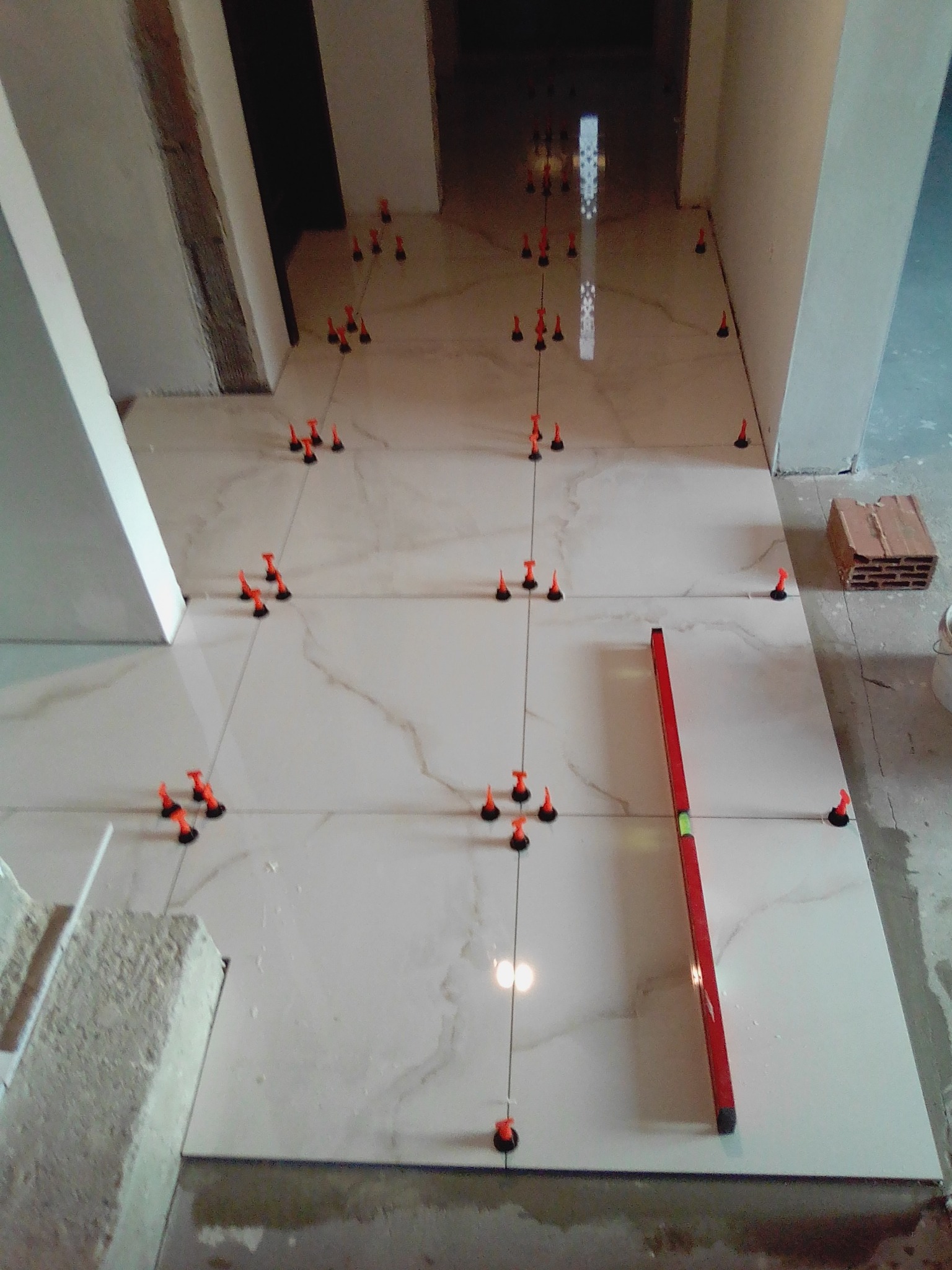 Układanie jasnych, marmurkowych płytek podłogowych z użyciem systemu poziomowania z pomarańczowymi klipsami i czerwonej poziomicy w długim korytarzu.