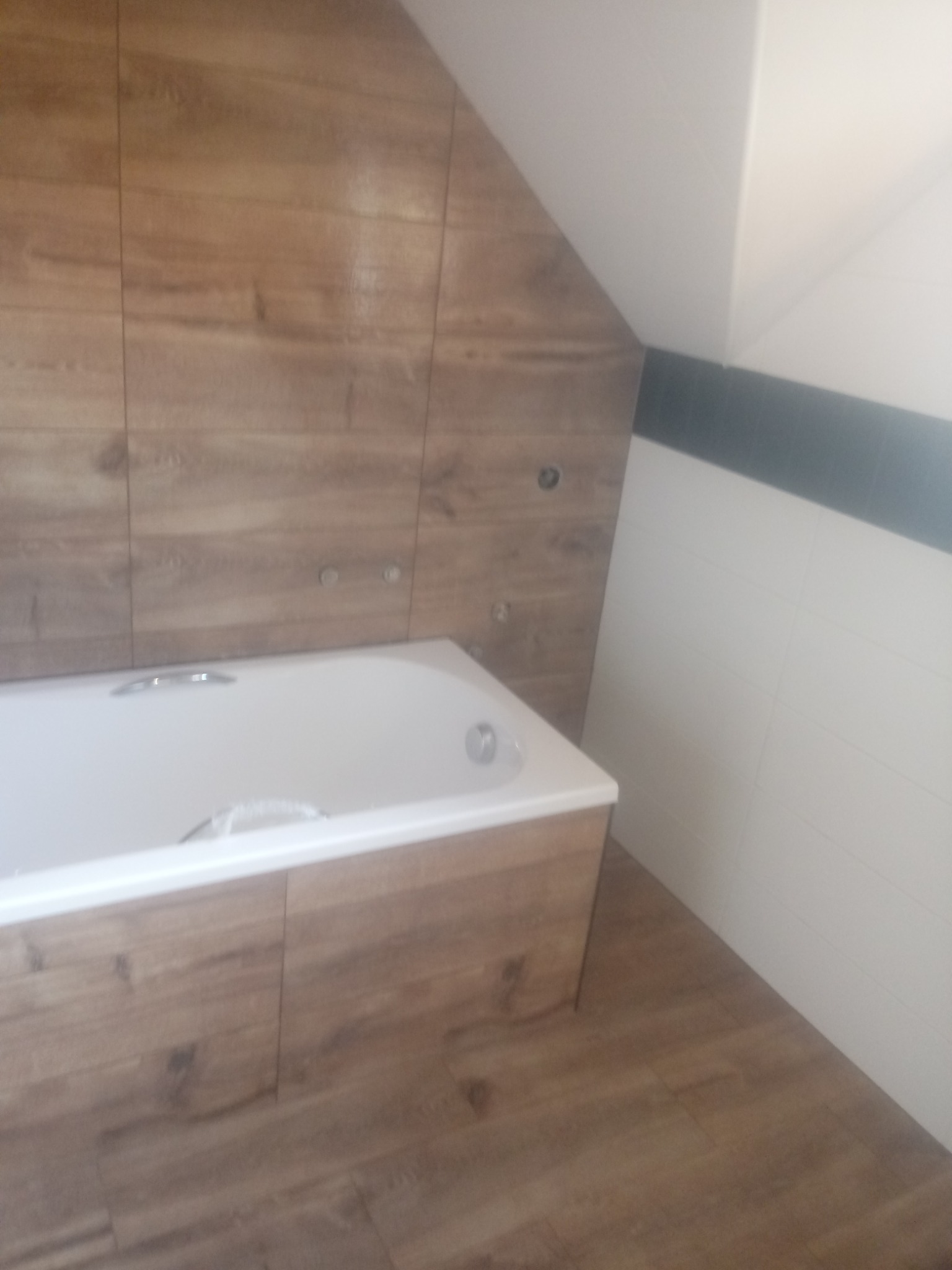 Biała wanna obudowana panelami drewnopodobnymi, z drewnopodobnymi płytkami na ścianie pod skosem dachu, widoczne chromowane elementy armatury, białe płytki na sąsiedniej ścianie oddzielone czarnym...