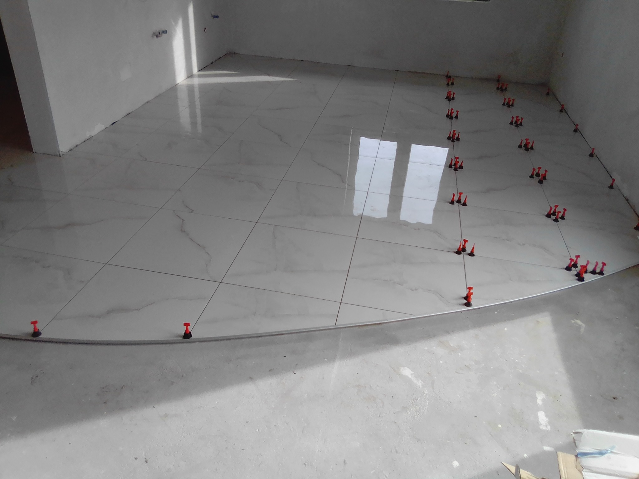 Układanie białych płytek podłogowych z marmurowym wzorem, z użyciem czerwonych klipsów poziomujących, widoczne odbicie okna na powierzchni płytek.