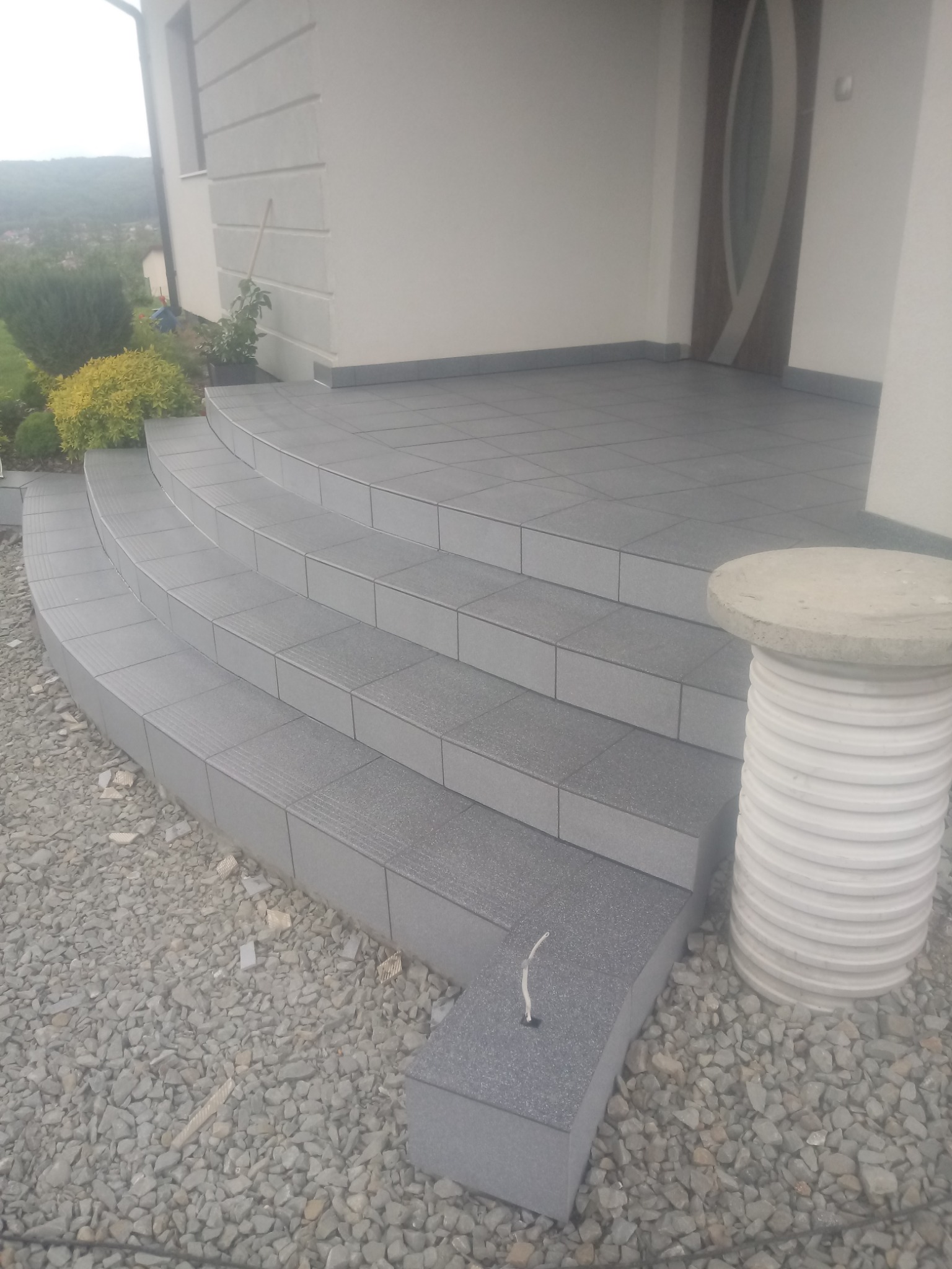 Wykończone płytkami schody wejściowe do domu z zaokrąglonymi stopniami, otoczone kamiennym żwirem, z widoczną kolumną dekoracyjną i fragmentem elewacji.