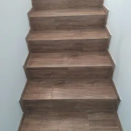 Schody wyłożone brązowymi płytkami imitującymi drewno, z widocznymi fugami i białymi ścianami po bokach.