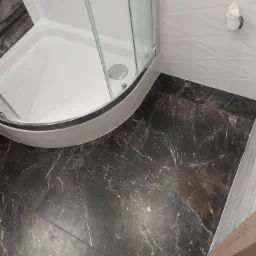 Narożnik kabiny prysznicowej z brodzikiem i nową, ciemną podłogą imitującą marmur, białe ściany z delikatnym wzorem geometrycznym i widoczną instalacją wodną.