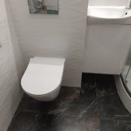 Nowoczesna toaleta wisząca z białą miską i chromowanym przyciskiem spłukującym na tle białych płytek z geometrycznym wzorem, uzupełniona umywalką z szafką i ciemną marmurkową podłogą.