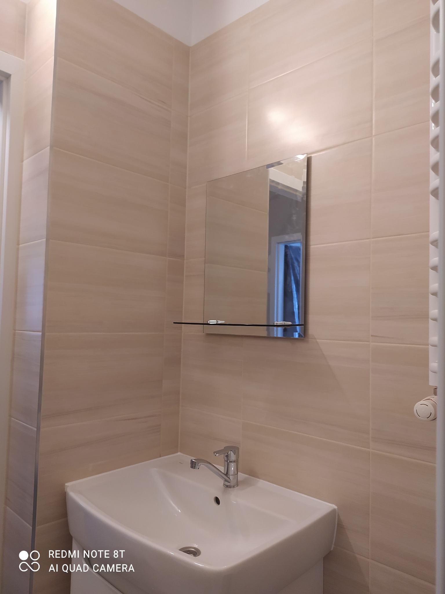 Jasna łazienka po remoncie: beżowe płytki imitujące kamień na ścianach, biała umywalka z chromowaną baterią, lustro z półką i grzejnik drabinkowy.