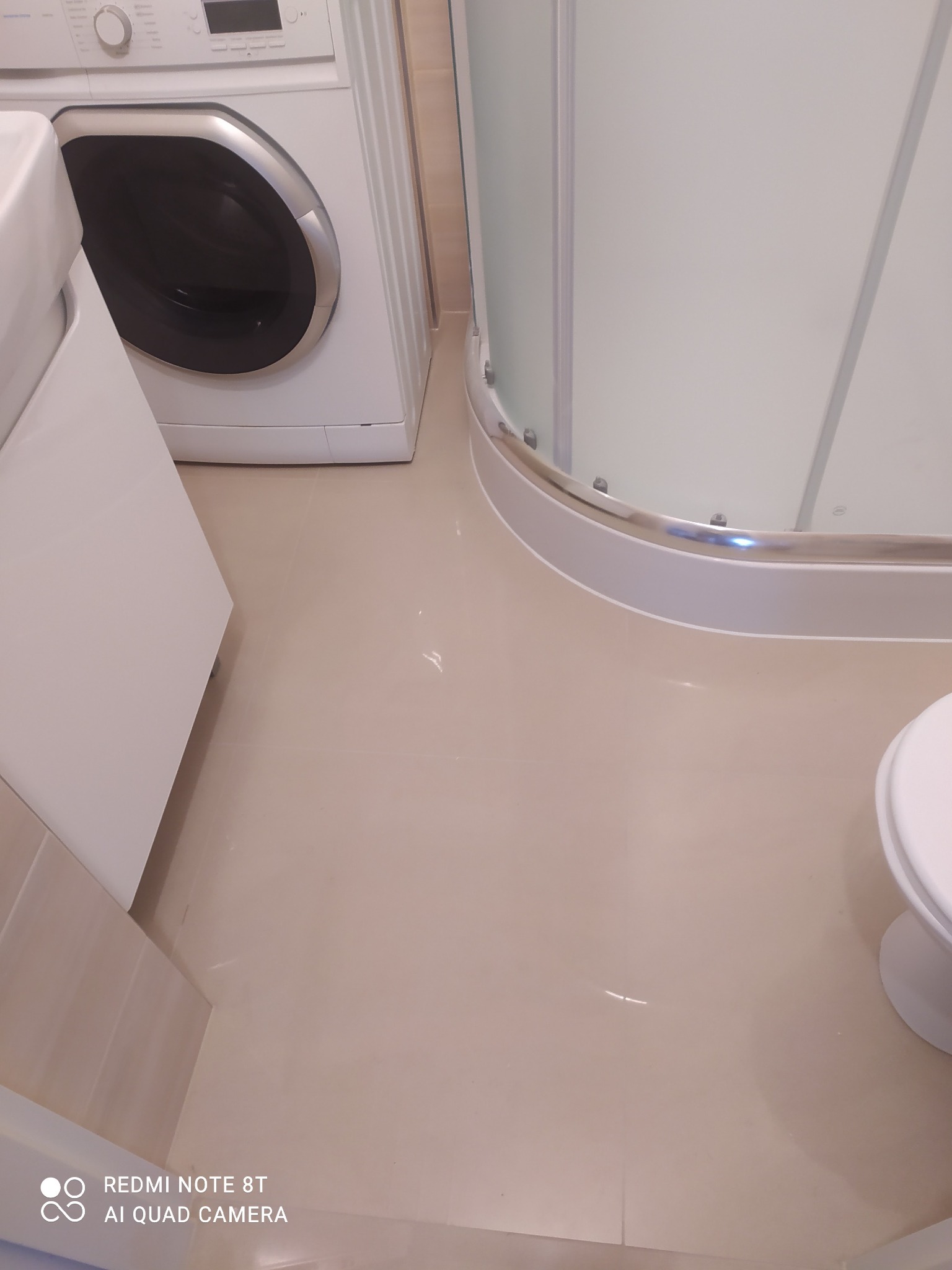 Beżowe płytki podłogowe w łazience, widoczna kabina prysznicowa z chromowanymi elementami, pralka i fragment szafki pod umywalkę.