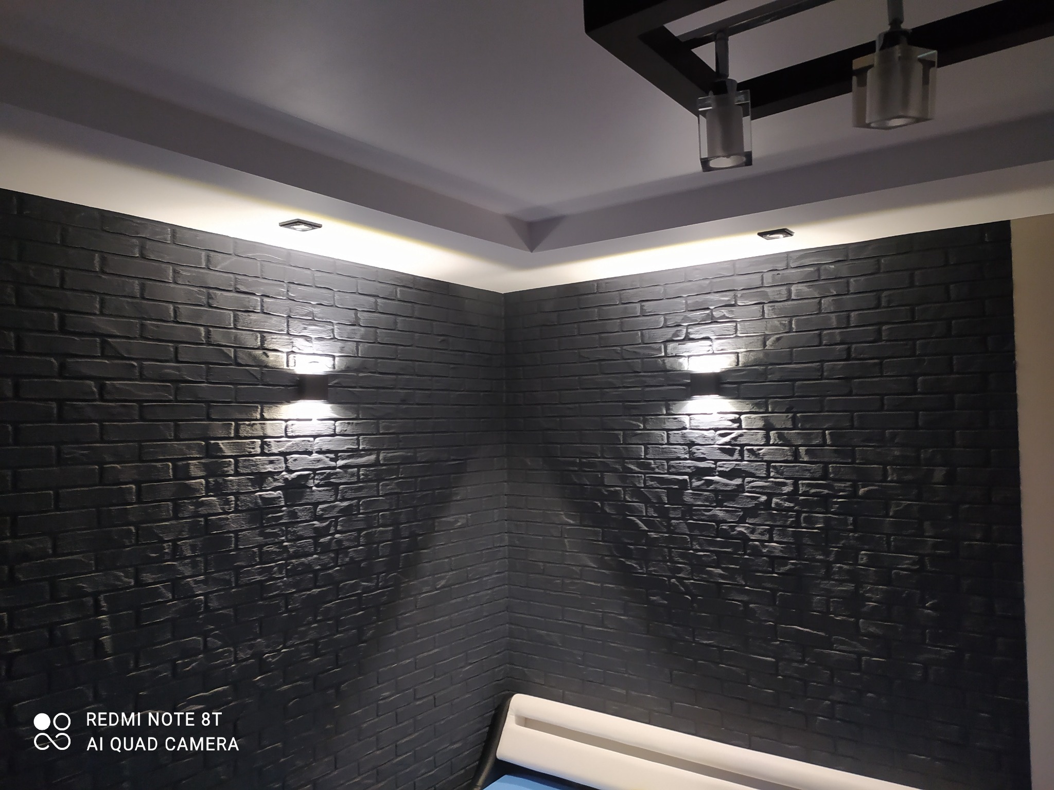 Ściana wykończona czarną cegłą dekoracyjną z oświetleniem punktowym i ledowym, widoczny fragment sufitu z lampami.