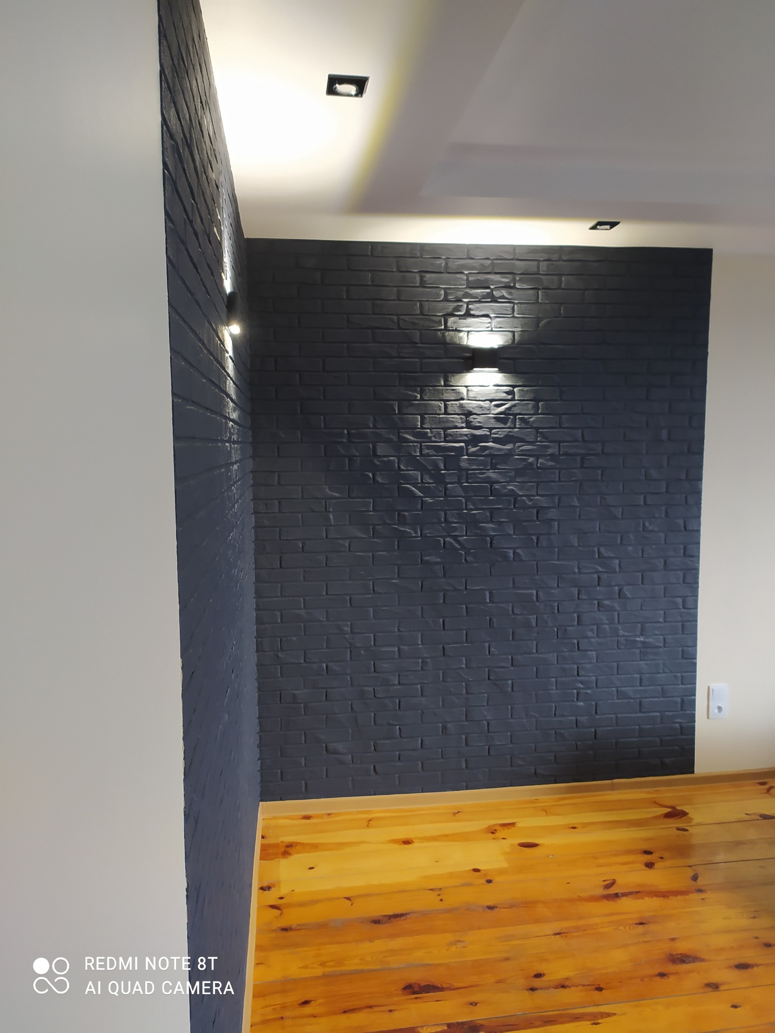 Ściana z cegły pomalowanej na ciemny grafit, oświetlona dwoma kinkietami, z widoczną podłogą z jasnego drewna i białymi ścianami po bokach, oświetlenie punktowe na suficie.