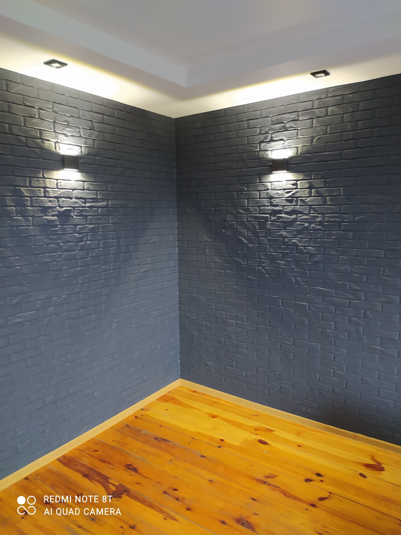 Wnętrze pokoju z pomalowaną na szaro ścianą imitującą cegłę, oświetloną przez dwie lampy ścienne i drewnianą podłogą.