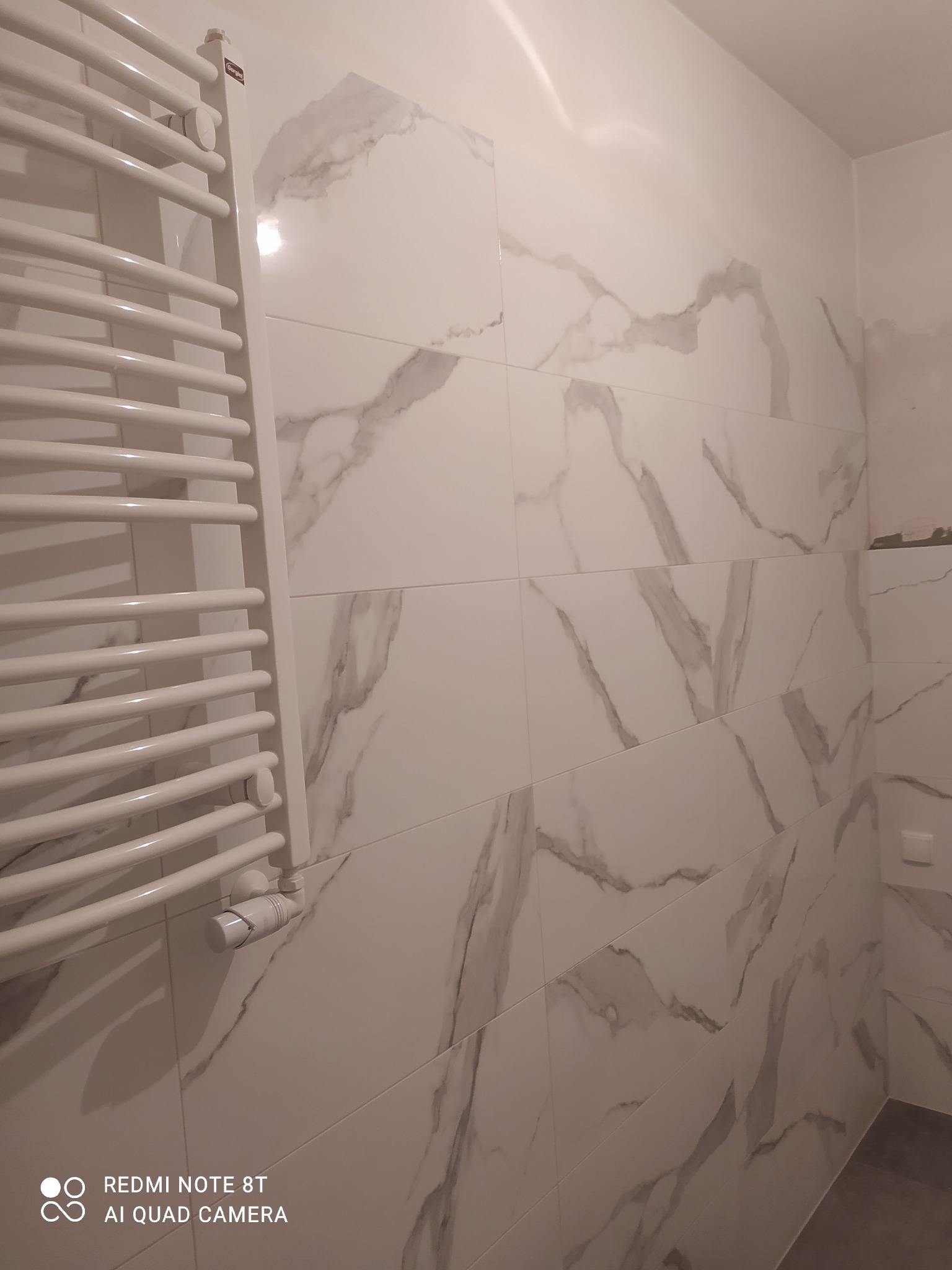 Ściana wyłożona białymi płytkami imitującymi marmur w łazience, z grzejnikiem drabinkowym po lewej stronie i fragmentem surowej ściany w prawym górnym rogu.
