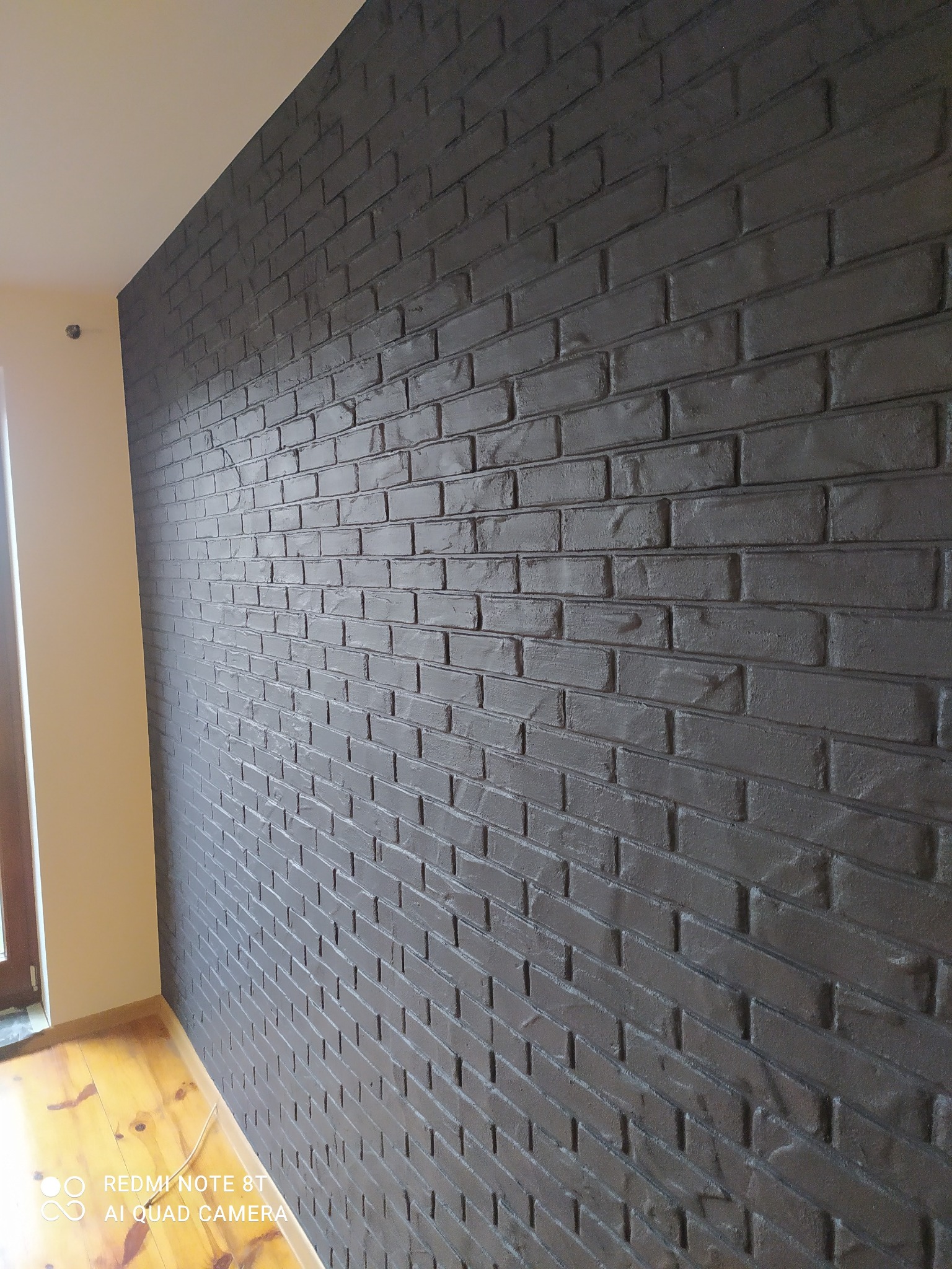 Ściana wykończona w stylu imitacji ciemnej cegły, z widoczną fakturą i fugami, oświetlona naturalnym światłem wpadającym z okna po lewej stronie, na podłodze widoczne deski parkietu.