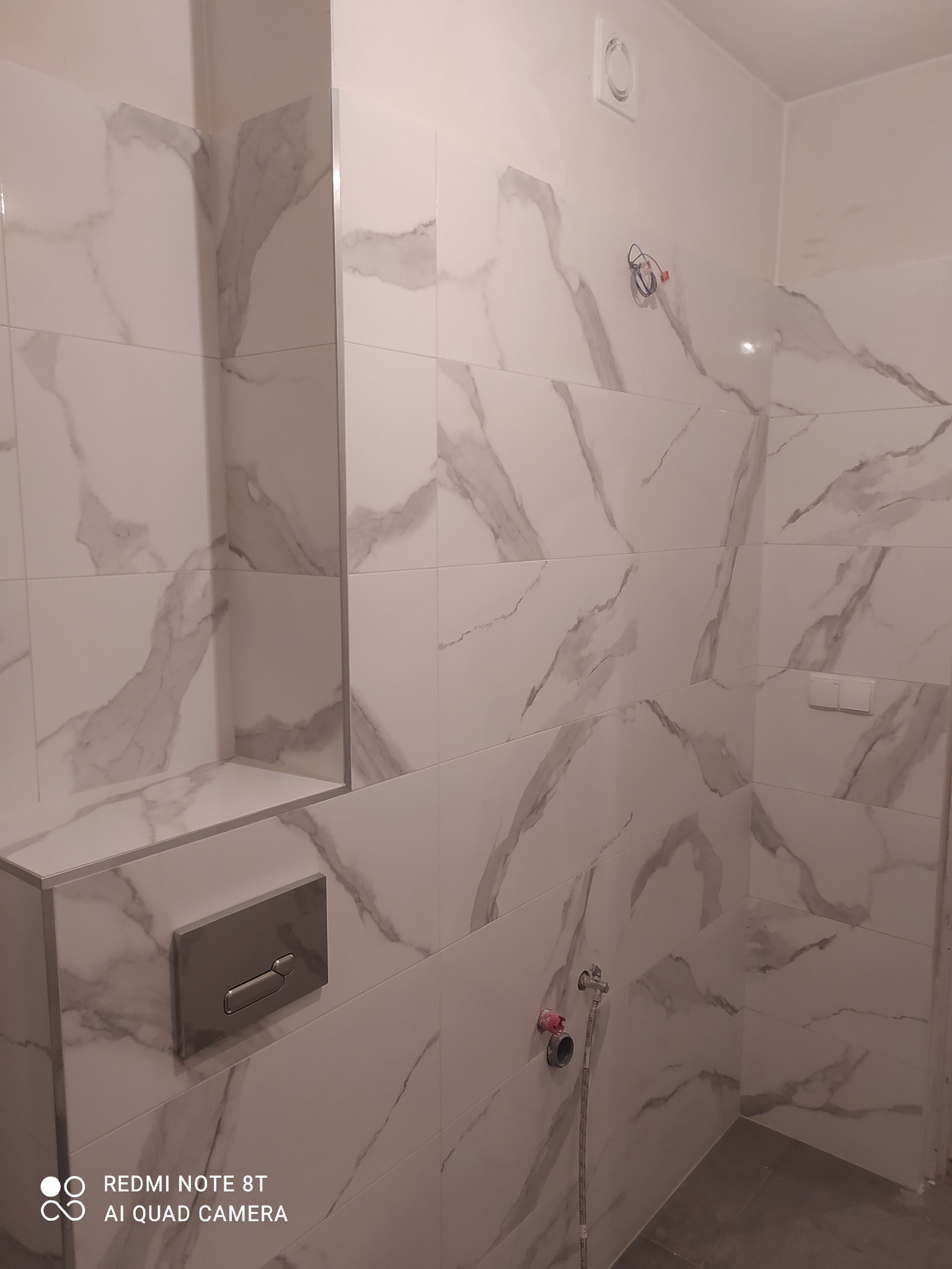 Łazienka wykończona białymi płytkami imitującymi marmur, widoczna zabudowa stelaża WC z chromowanym przyciskiem oraz wnęka z półką wyłożona tymi samymi płytkami, instalacja wodna w trakcie montażu.