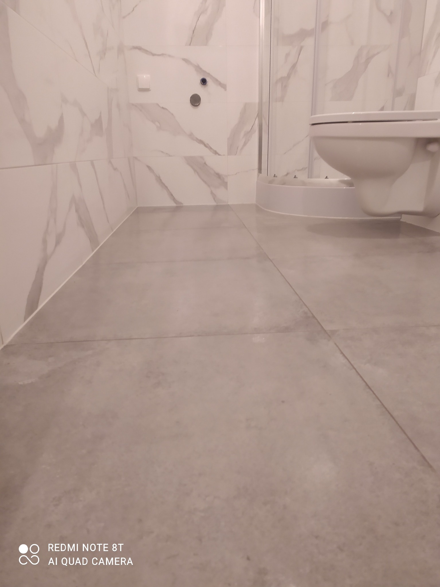 Nowoczesna łazienka po remoncie: szare płytki podłogowe, marmurowe płytki ścienne, kabina prysznicowa i toaleta w odcieniach bieli.