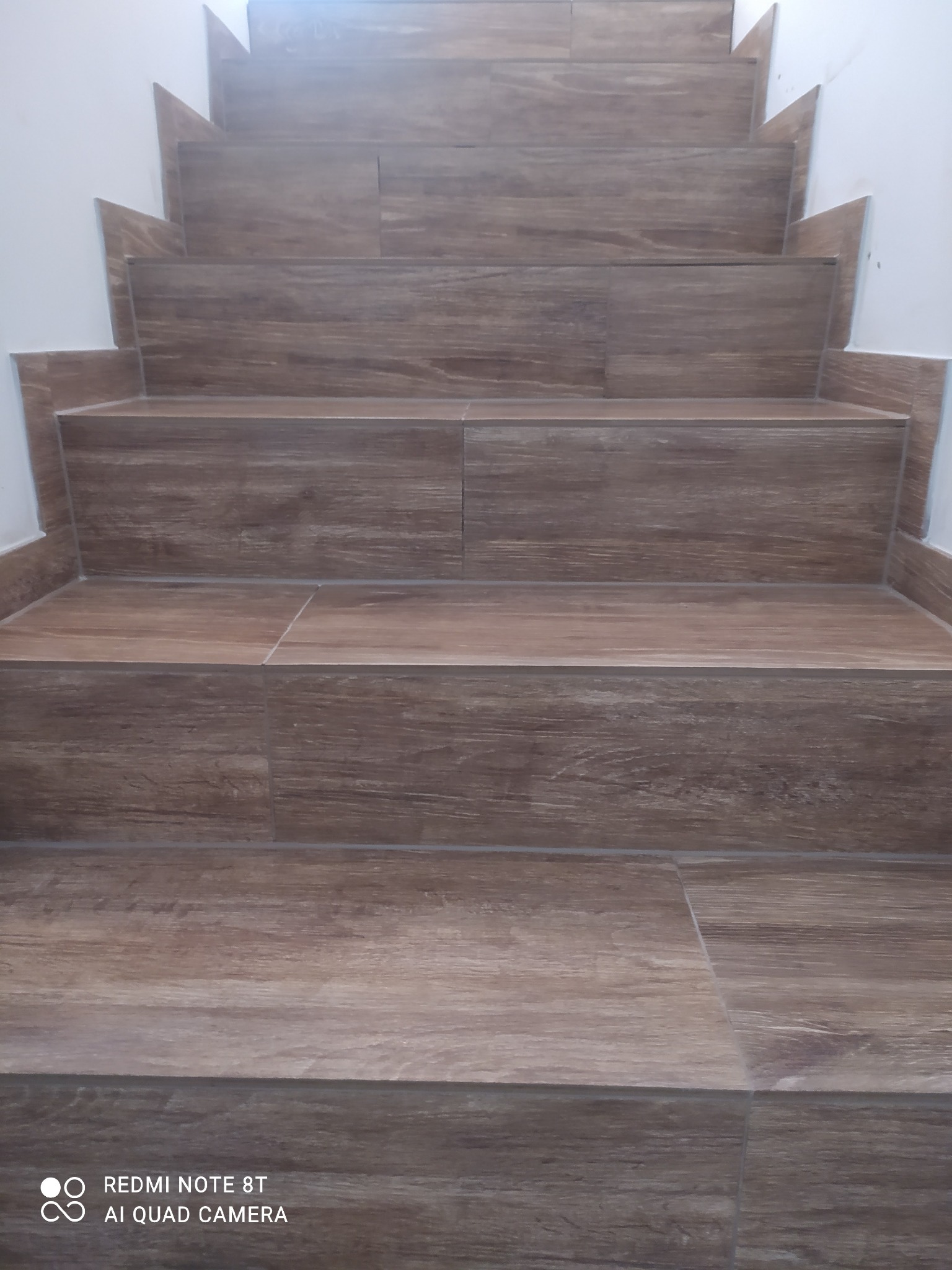 Schody wyłożone brązowymi płytkami imitującymi drewno, widok z dołu, białe ściany po bokach, widoczne fugi.