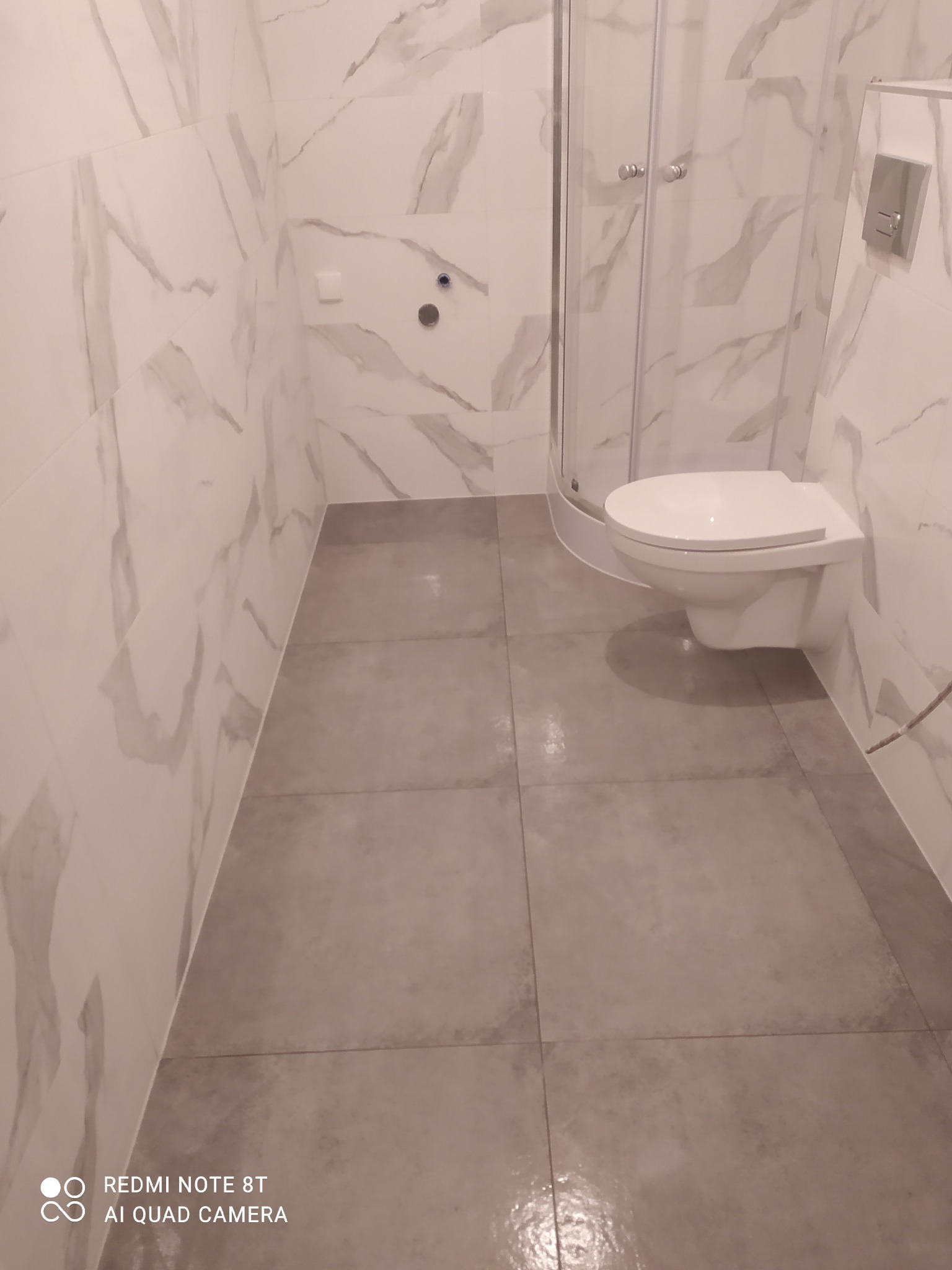 Wykończona łazienka z biało-szarymi marmurkowymi płytkami na ścianach, szarą podłogą z płytek, kabiną prysznicową z przezroczystego szkła i podwieszaną toaletą.