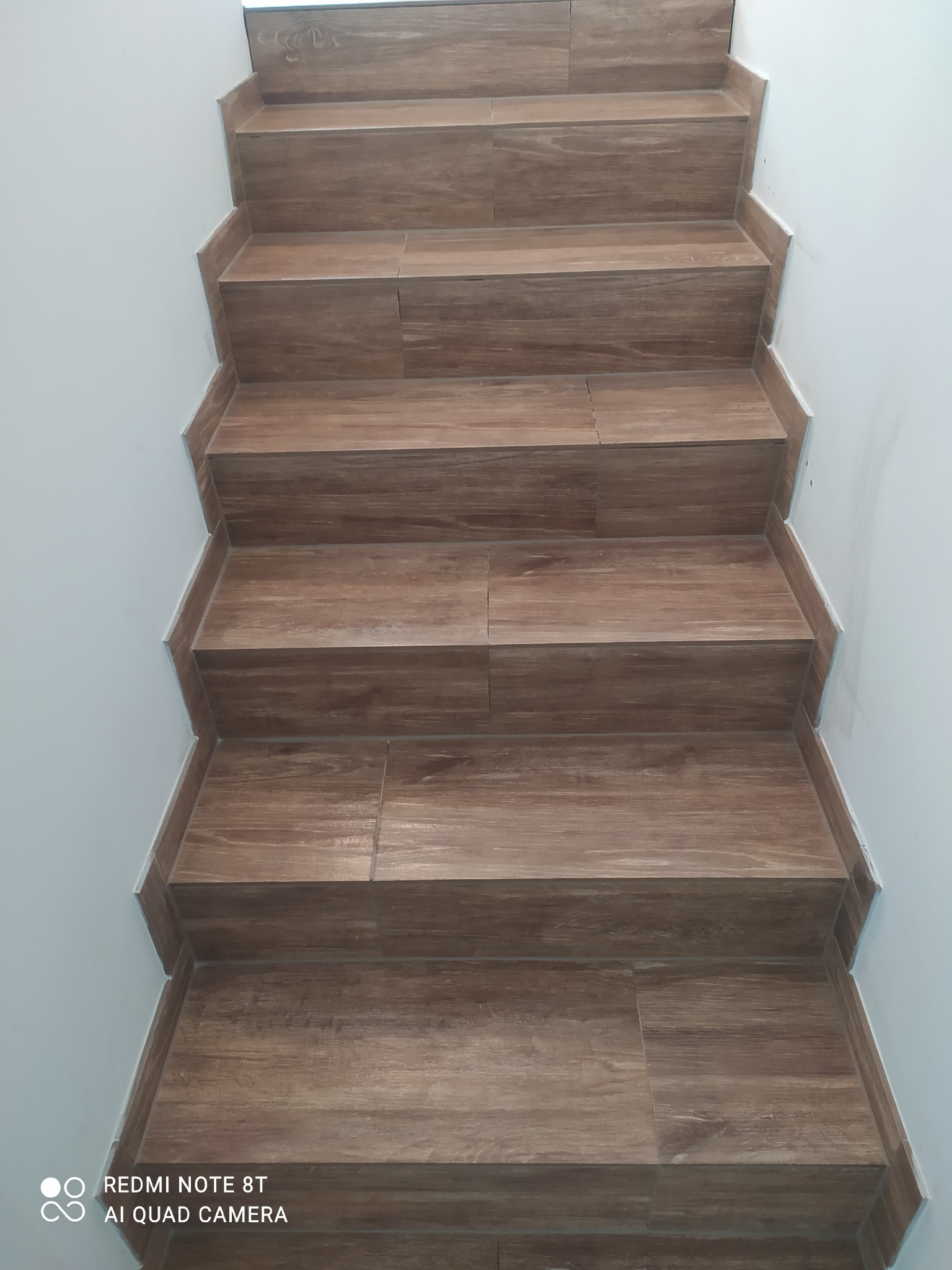 Schody wyłożone brązowymi płytkami imitującymi drewno, z widocznymi fugami i białymi ścianami po bokach.