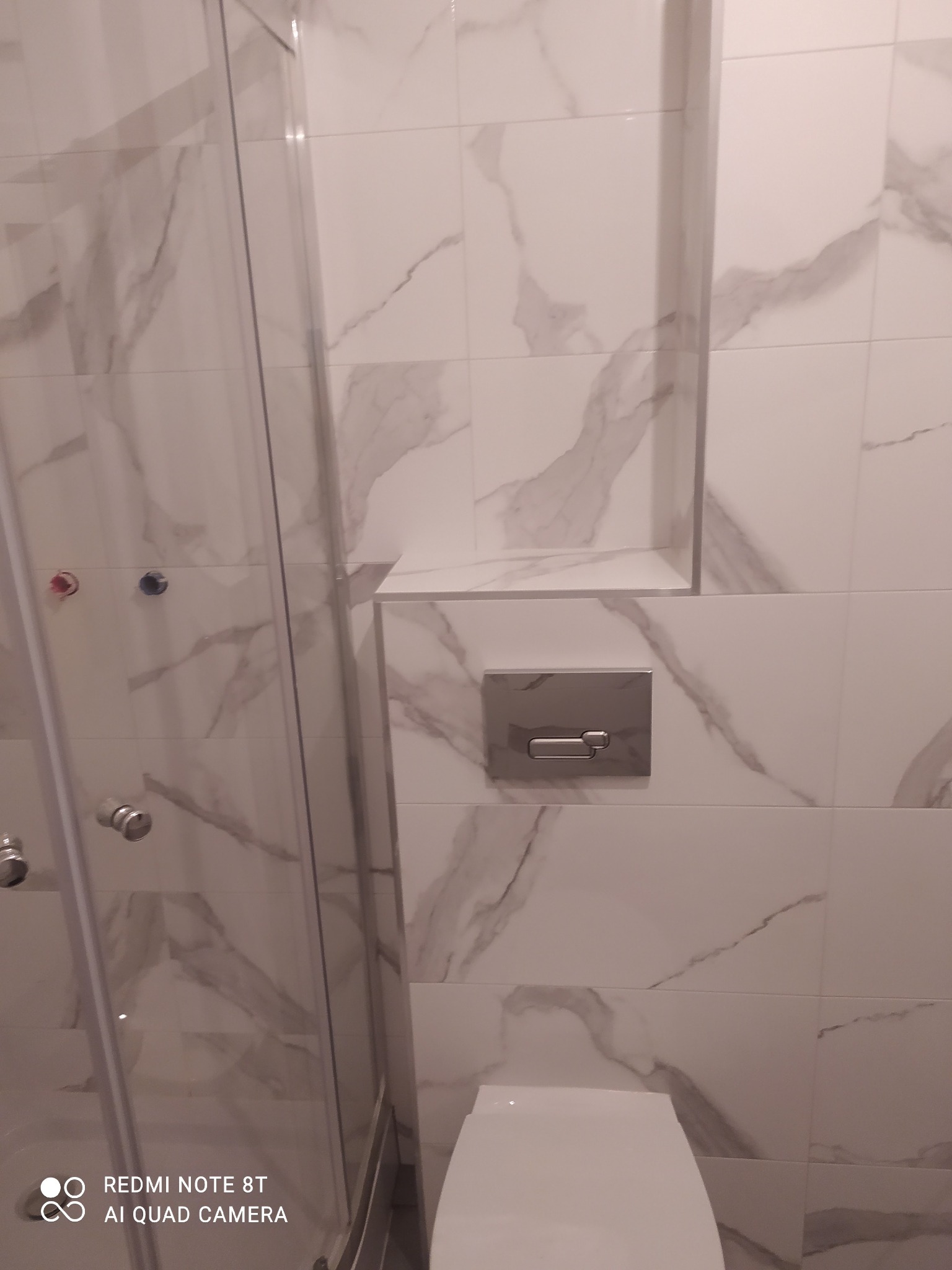 Biała łazienka z marmurowymi płytkami, widoczna kabina prysznicowa i zabudowana toaleta z chromowanym przyciskiem spłukującym, dodatkowo w ścianie wnęka wykończona tymi samymi płytkami.