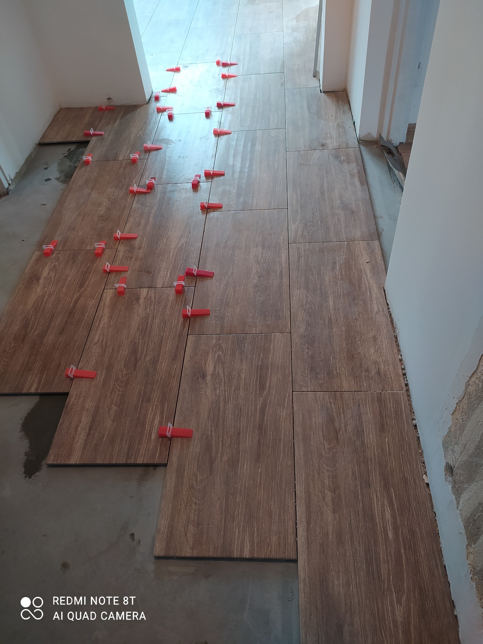 Układanie brązowych płytek podłogowych imitujących drewno w korytarzu, z użyciem czerwonych klipsów poziomujących, widoczne niedokończone fragmenty przy ścianach.