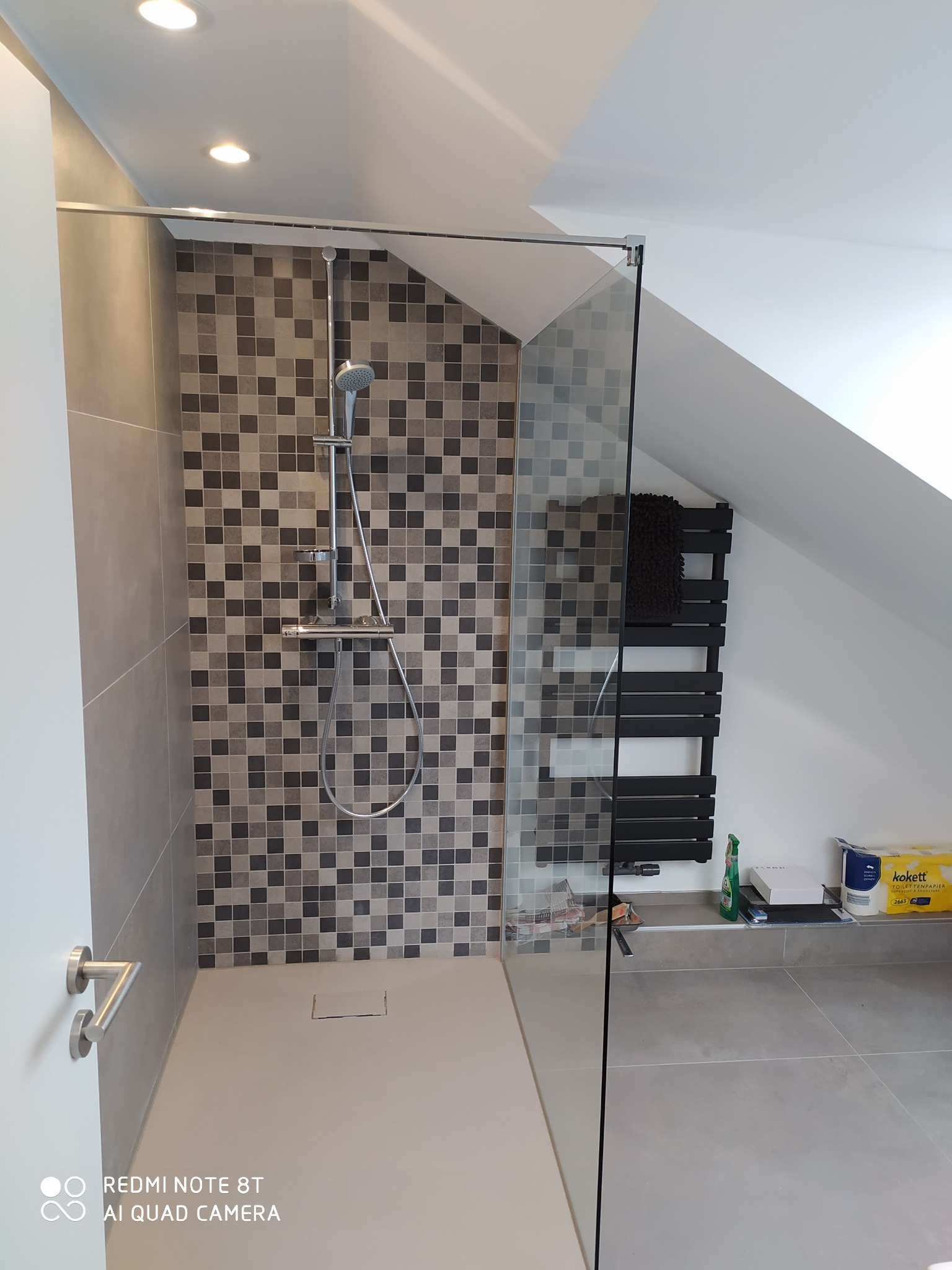 Nowoczesna kabina prysznicowa w łazience na poddaszu, wykończona mozaiką w odcieniach szarości, z odpływem liniowym i szklaną ścianką; obok grzejnik drabinkowy w kolorze czarnym.