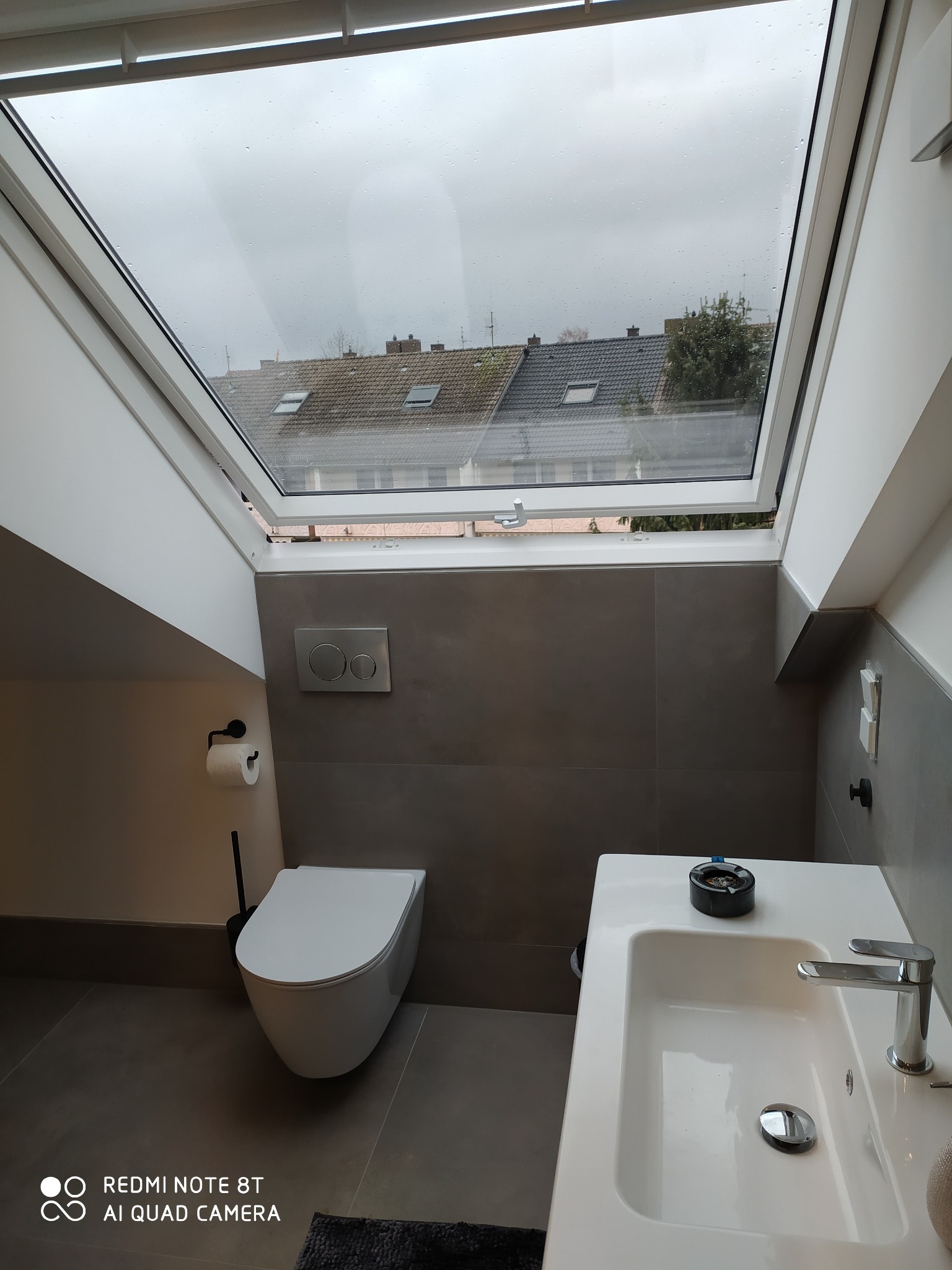 Nowoczesna łazienka na poddaszu z oknem dachowym, szare płytki ścienne i podłogowe, wisząca toaleta, biała umywalka z chromowaną baterią, widok na dachy sąsiednich budynków przez okno.