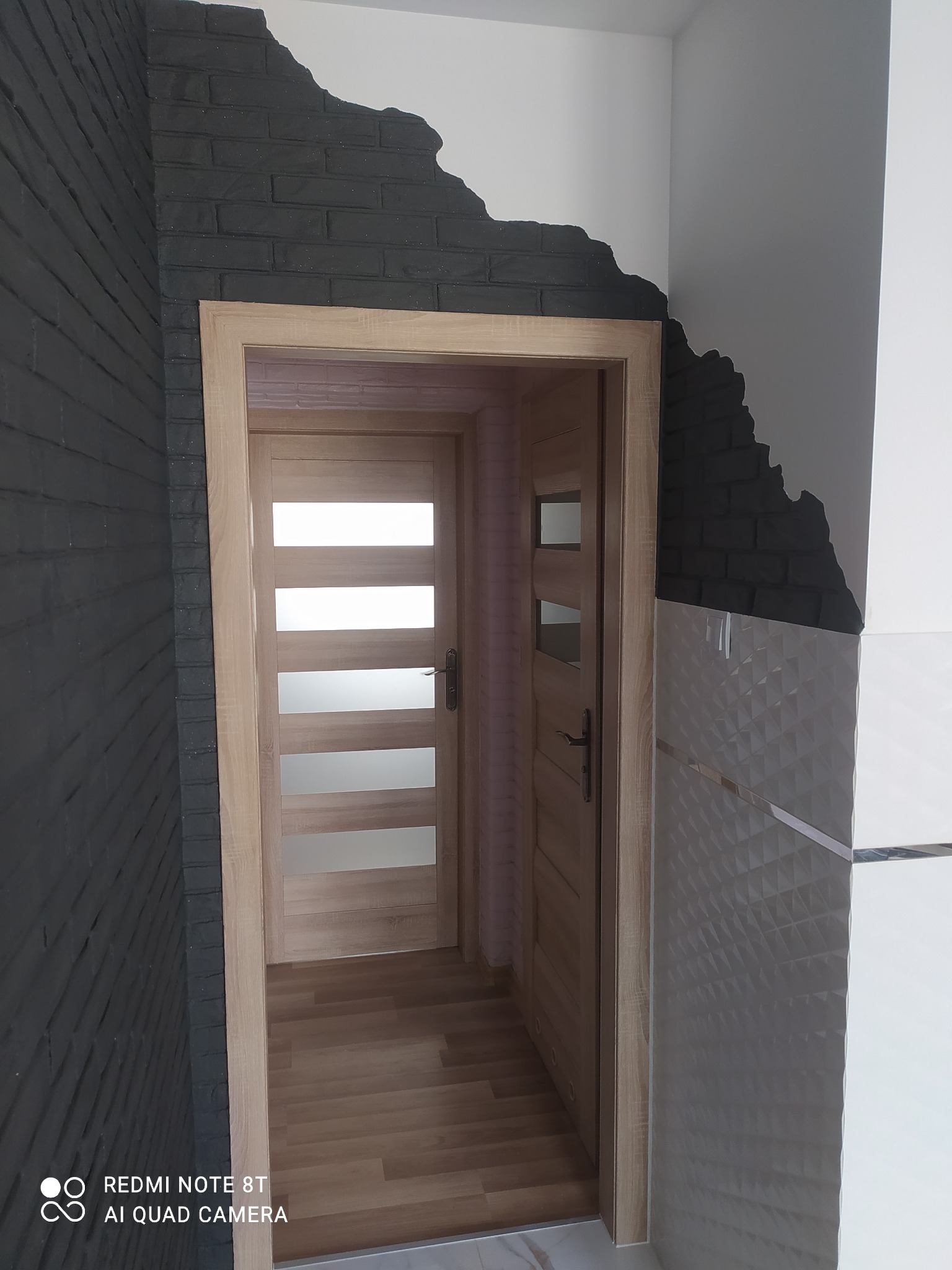 Wykończenie wnętrza z efektem surowej cegły pomalowanej na czarno przy futrynie drzwiowej i dekoracyjną płytką ścienną z geometrycznym wzorem, widok na drewniane drzwi wewnętrzne.