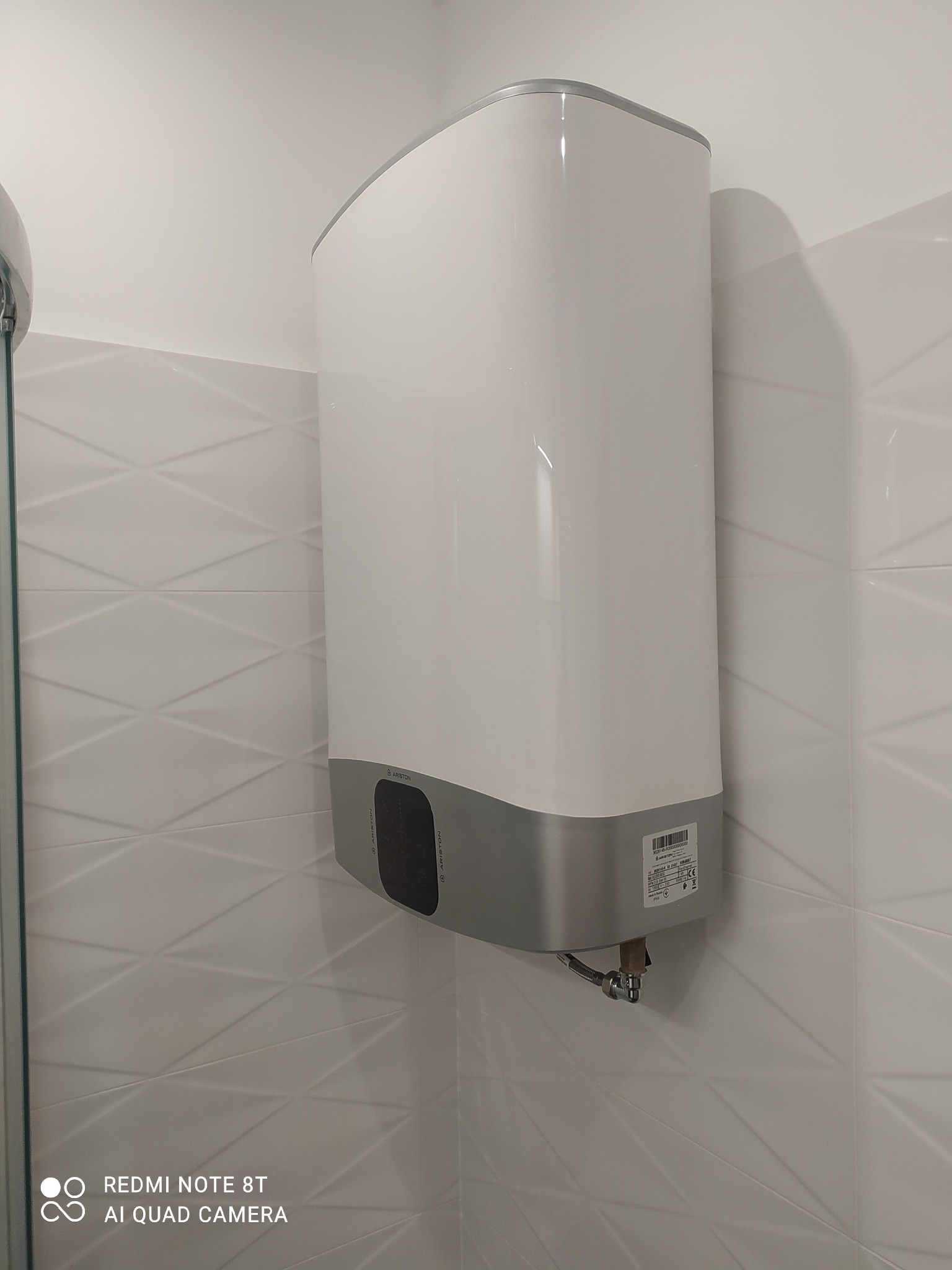Biały podgrzewacz wody marki Ariston zamontowany na ścianie łazienki wyłożonej białymi, trójwymiarowymi płytkami, z widocznym podłączeniem hydraulicznym.