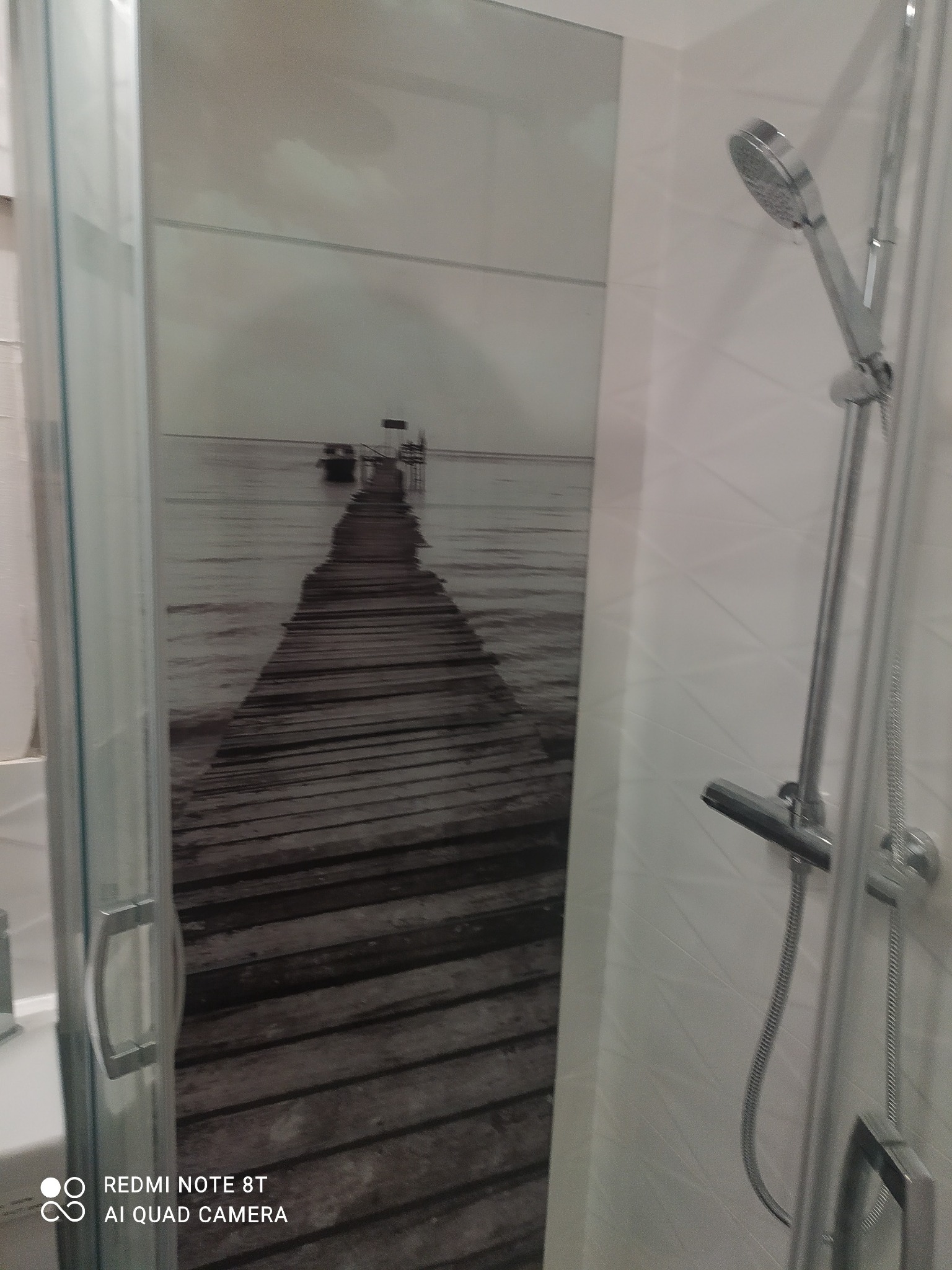 Kabina prysznicowa z panelem dekoracyjnym przedstawiającym molo i morze w odcieniach szarości, widoczna bateria i słuchawka prysznicowa na tle białej ściany z geometrycznym wzorem.