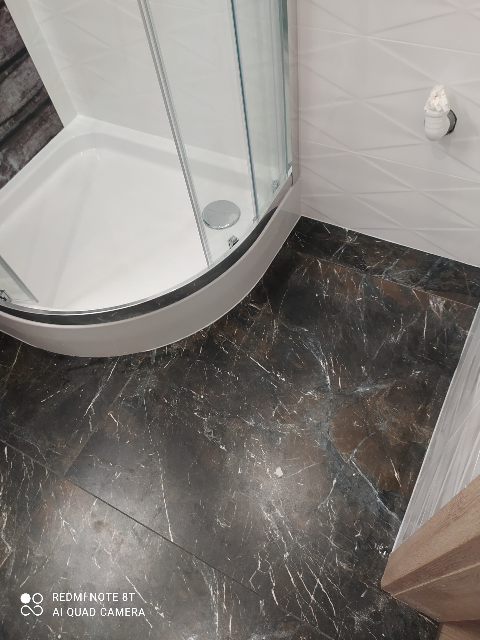 Narożnik kabiny prysznicowej z brodzikiem i nową, ciemną podłogą imitującą marmur, białe ściany z delikatnym wzorem geometrycznym i widoczną instalacją wodną.