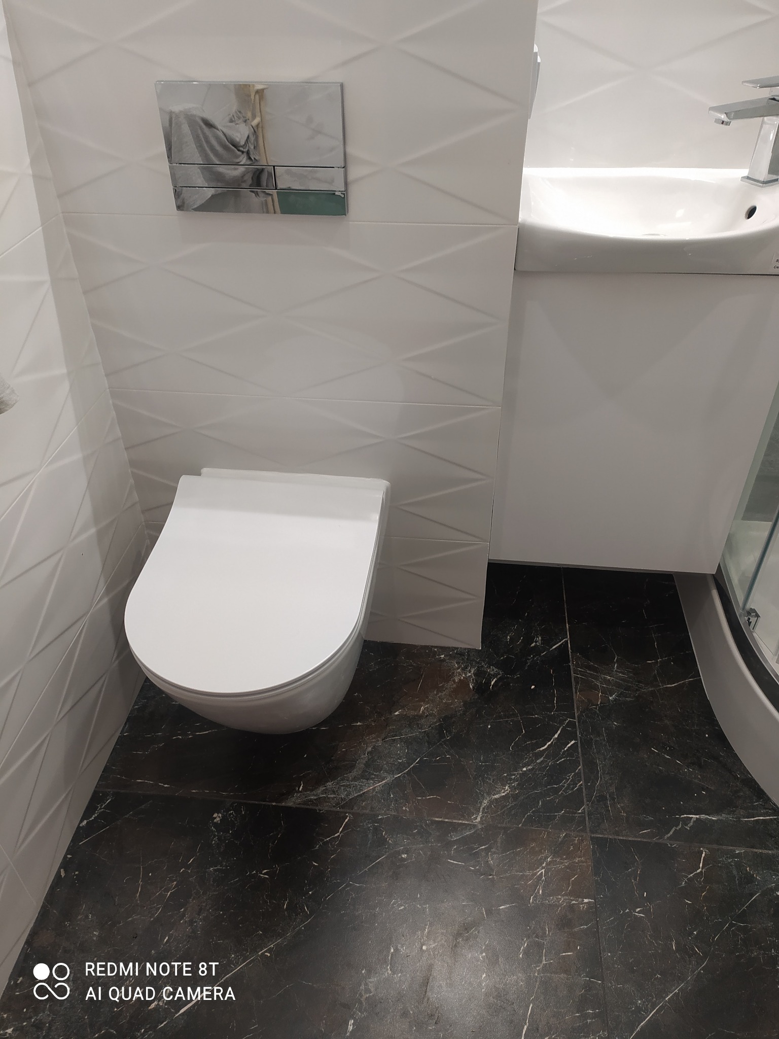 Nowoczesna toaleta wisząca z białą miską i chromowanym przyciskiem spłukującym na tle białych płytek z geometrycznym wzorem, uzupełniona umywalką z szafką i ciemną marmurkową podłogą.