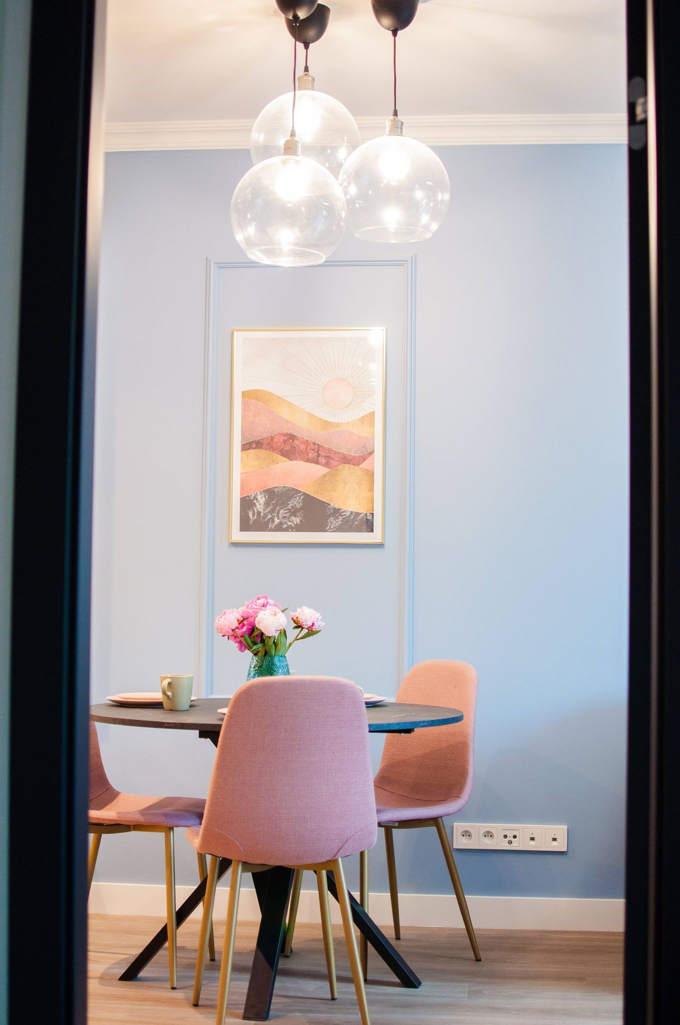 Jadalnia w pastelowych kolorach z okrągłym stołem, różowymi krzesłami na złotych nogach, kwiatami w turkusowym wazonie i abstrakcyjnym obrazem na ścianie w listwach.