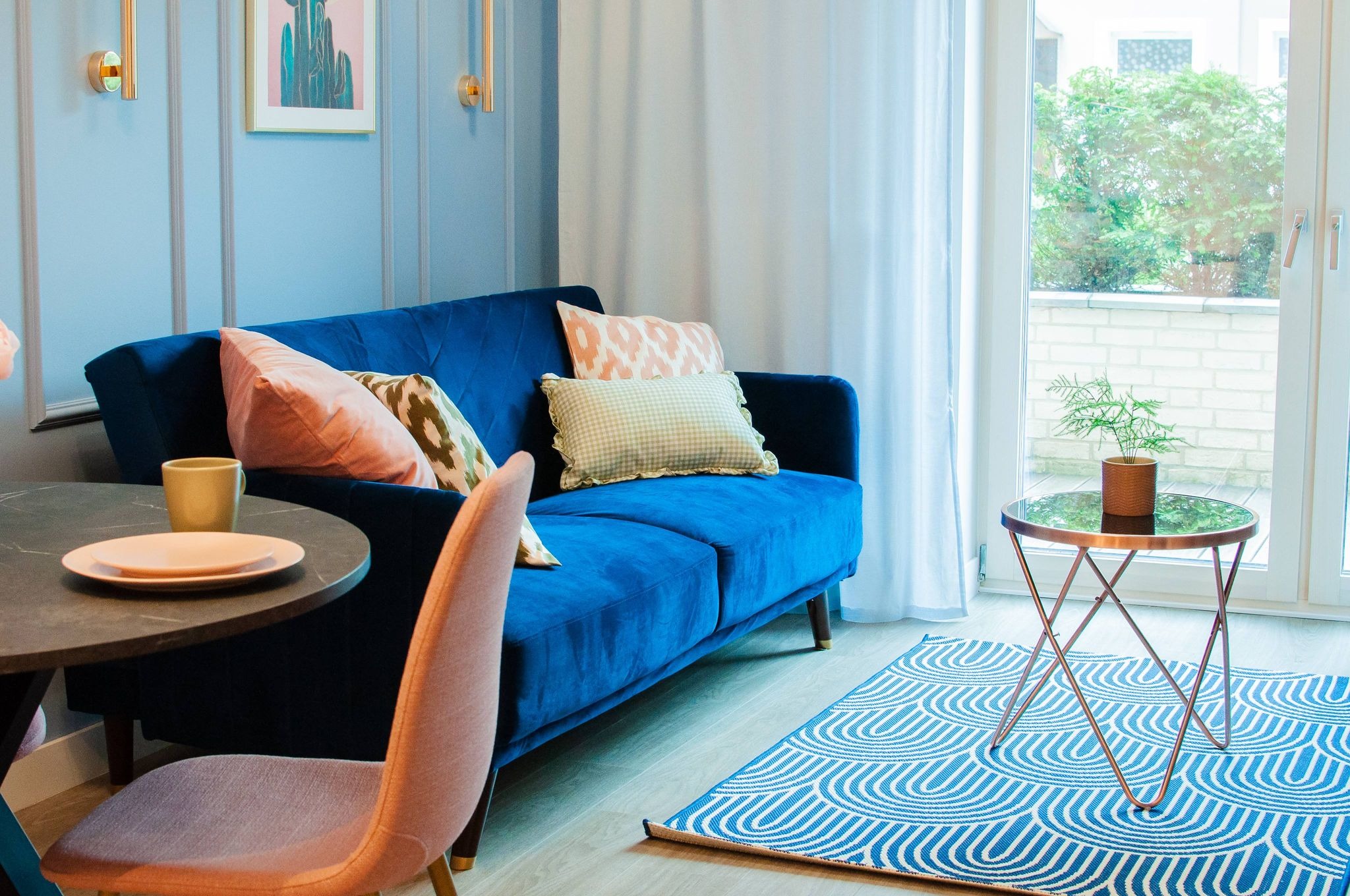 Niebieska welurowa sofa z poduszkami w pastelowych kolorach, okrągły stolik kawowy z rośliną w doniczce, okrągły stół z nakryciem, różowe krzesło, niebieski dywan w białe wzory, jasne ściany...