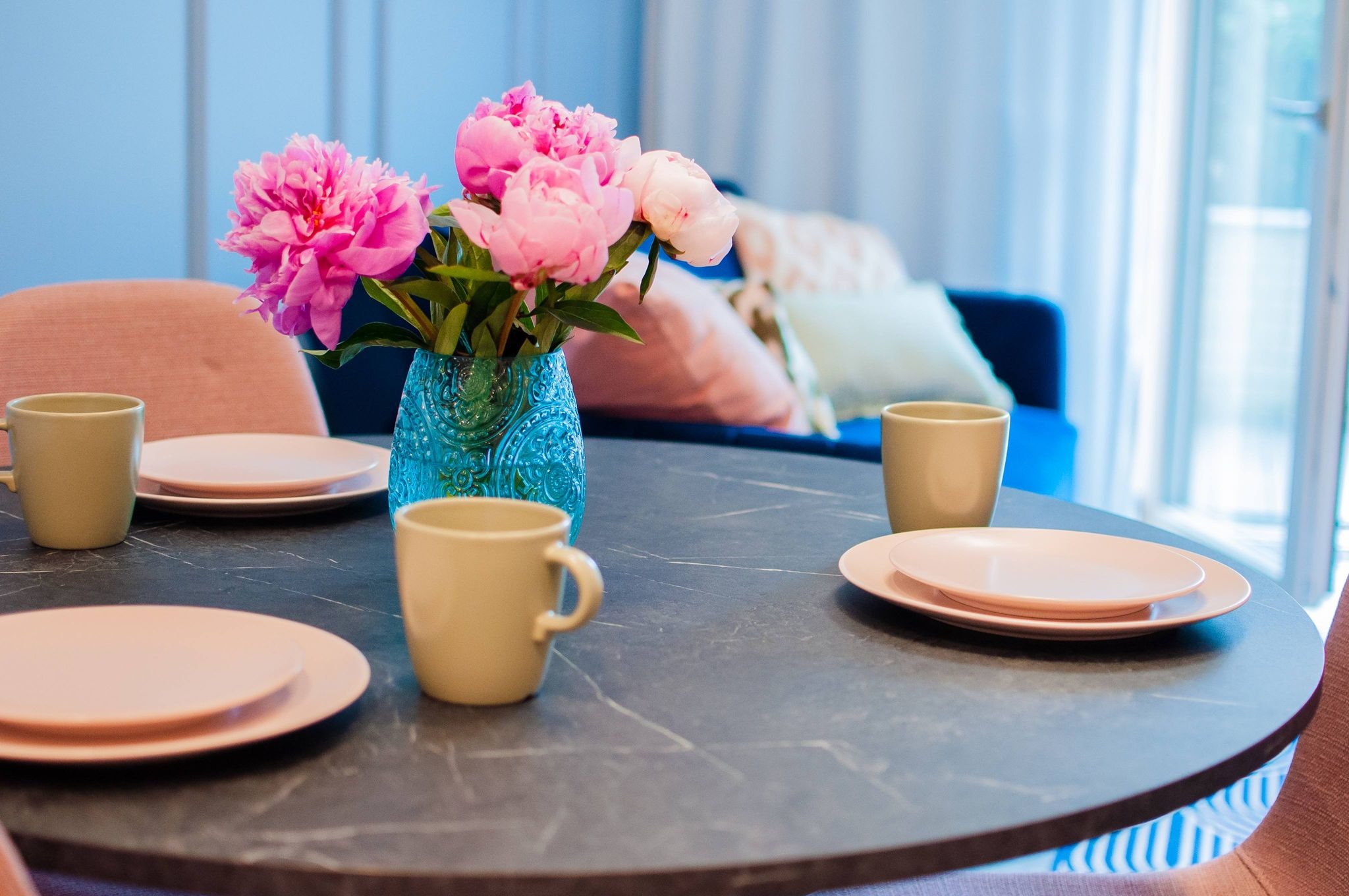 Stół z ciemnym blatem, nakryty różowymi talerzami i beżowymi kubkami, ozdobiony wazonem z różowymi piwoniami, w tle błękitna ściana i kanapa.