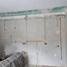 Ściana w trakcie prac remontowych z widocznymi przewodami elektrycznymi, gniazdkami i puszkami. Widoczne ślady po bruzdowaniu i mocowaniu przewodów.