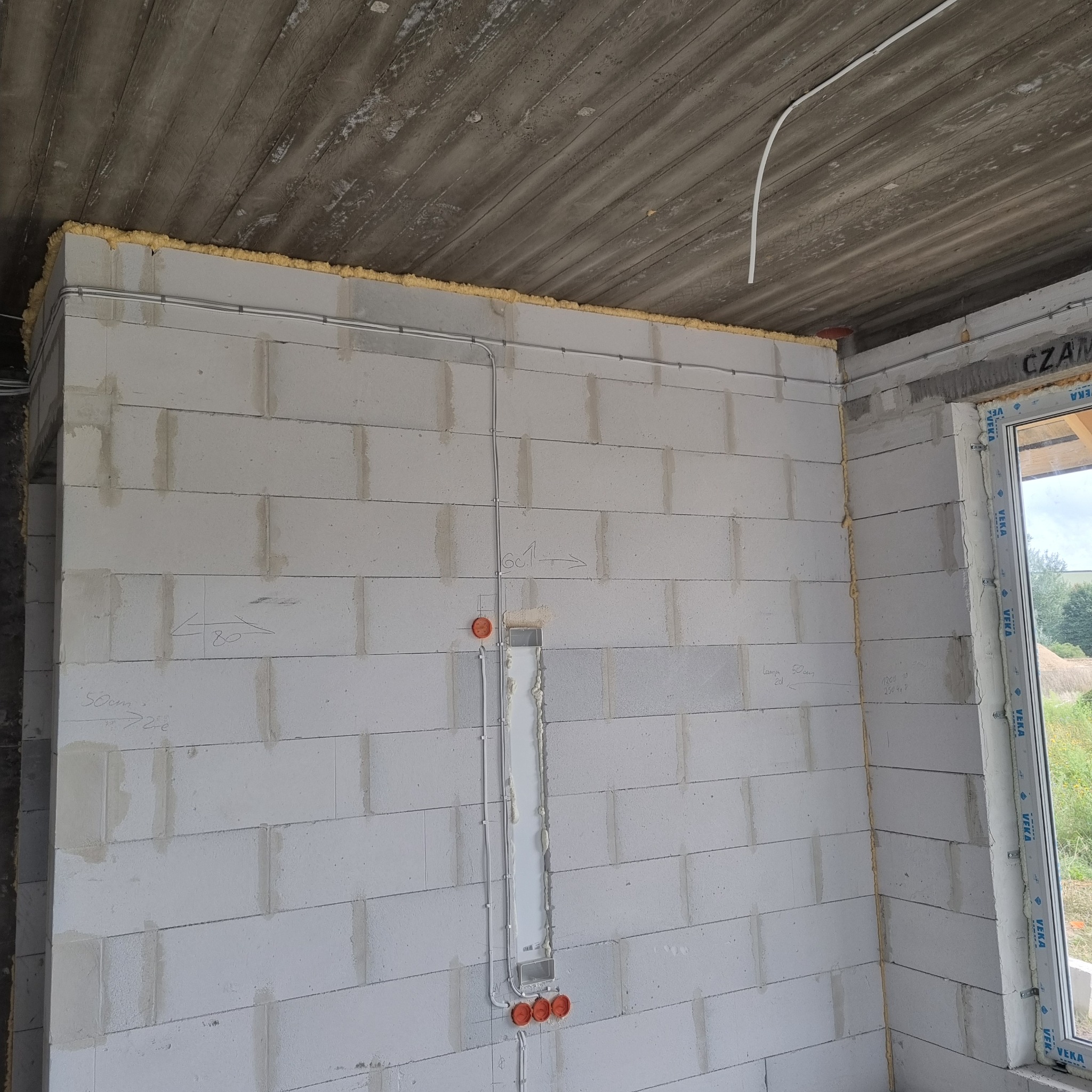 Ściana z instalacją elektryczną w surowym stanie: kable w peszlach, puszki podtynkowe, pianka montażowa przy oknie. Widoczne oznaczenia na ścianie.