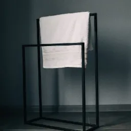 Minimalistyczny, czarny, metalowy stojak na ręczniki z białym, puszystym ręcznikiem na tle szarej ściany i podłogi z płytek.